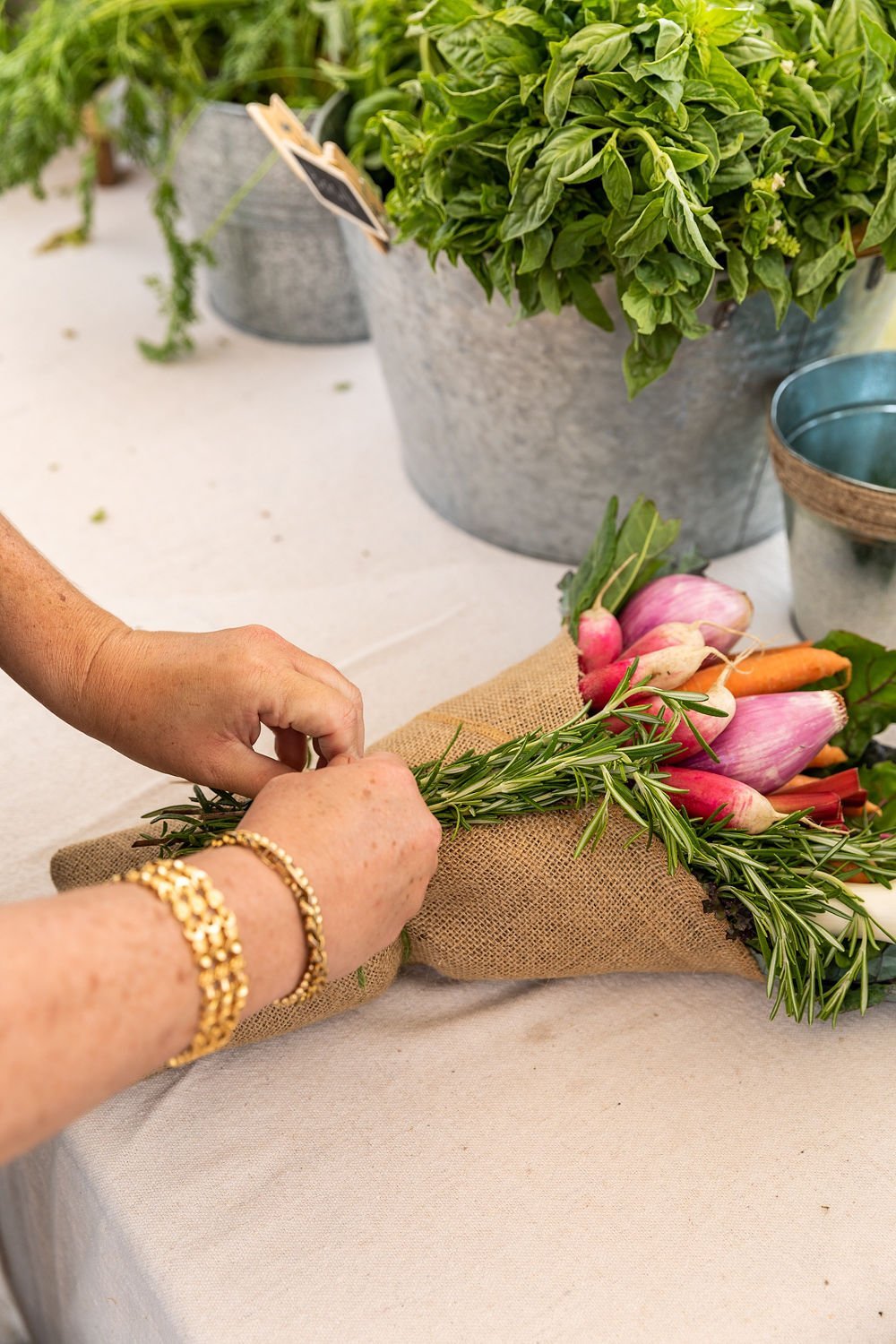 Person arranging a bouquet of pink radishes and sprigs of rosemary on a table with potted basil plants in the background.