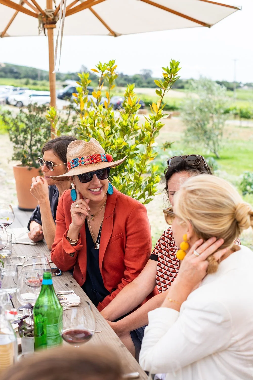 Group of four women enjoying outdoor meal at a wooden table, with one woman smiling and wearing a colorful hat and turquoise earrings, under an umbrella on a sunny day.