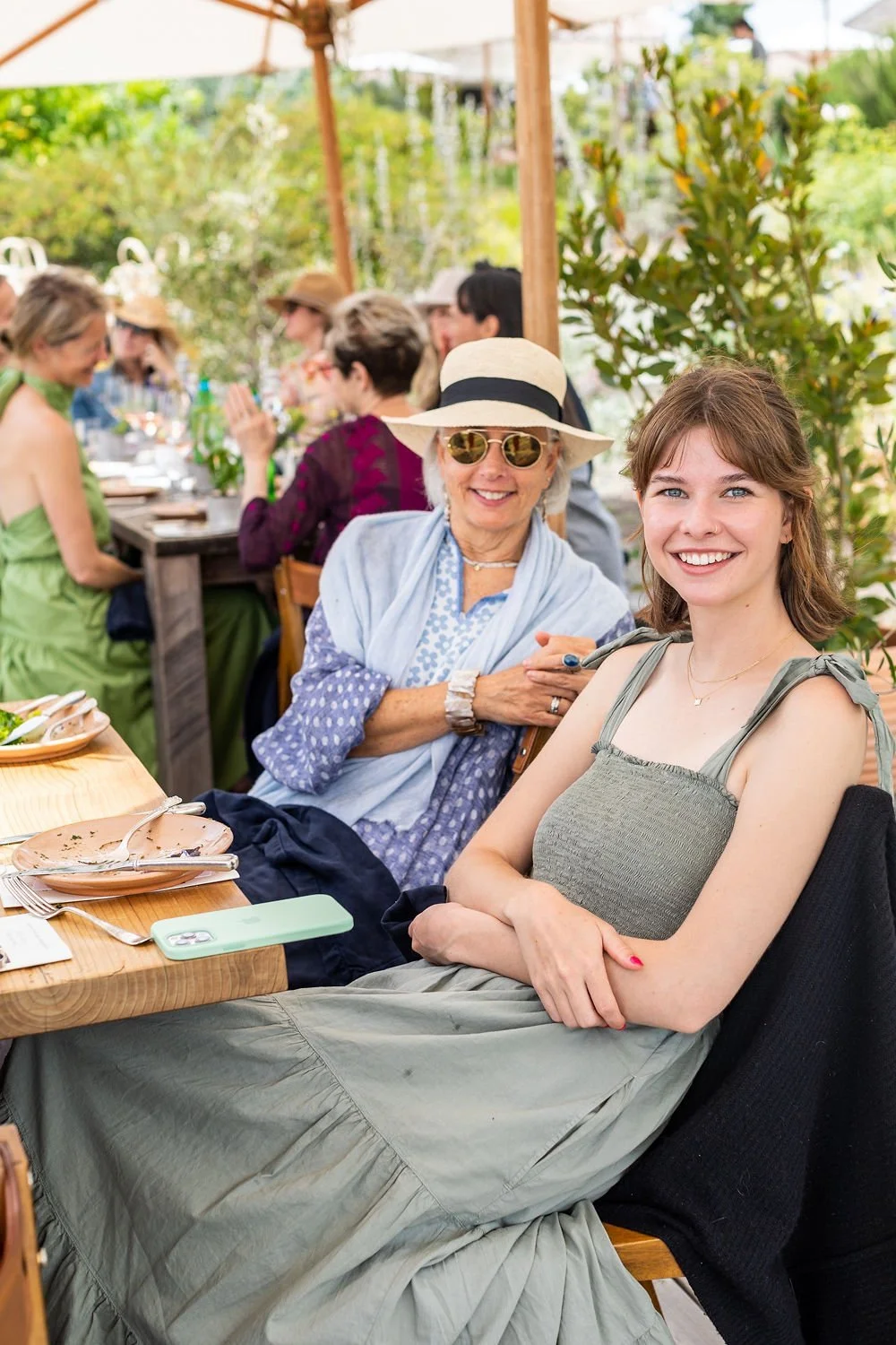 A group of women enjoying a meal outdoors at a sunny restaurant, smiling and socializing, with greenery and umbrellas in the background.