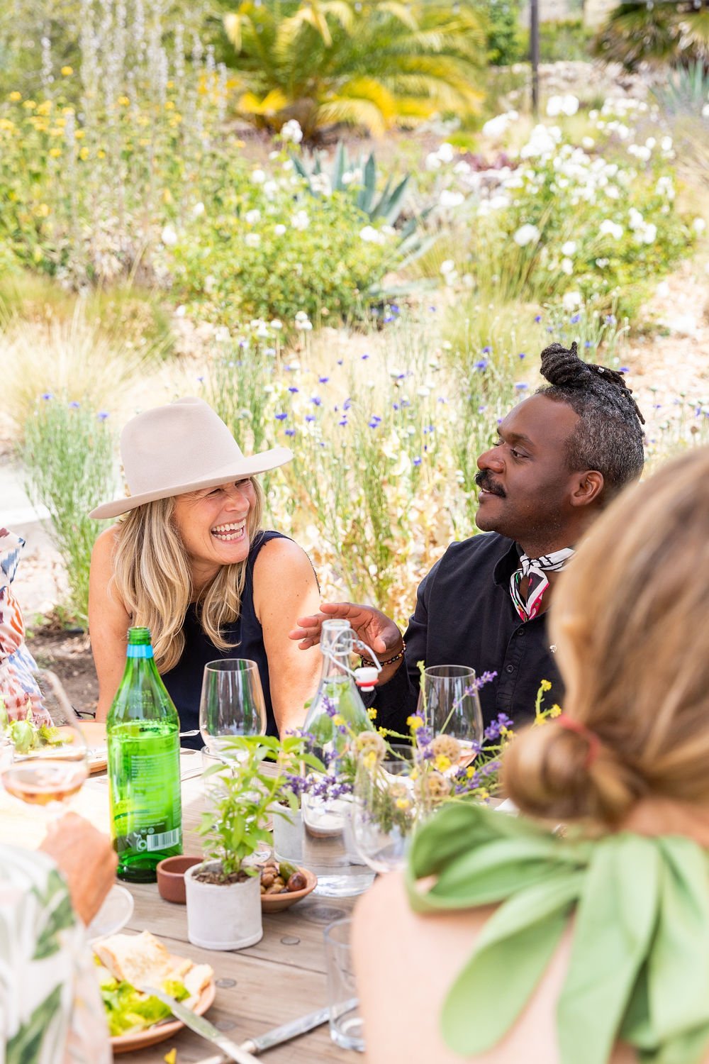 People enjoying a outdoor garden party, sitting at a table with drinks and flowers, surrounded by greenery and blooming flowers.