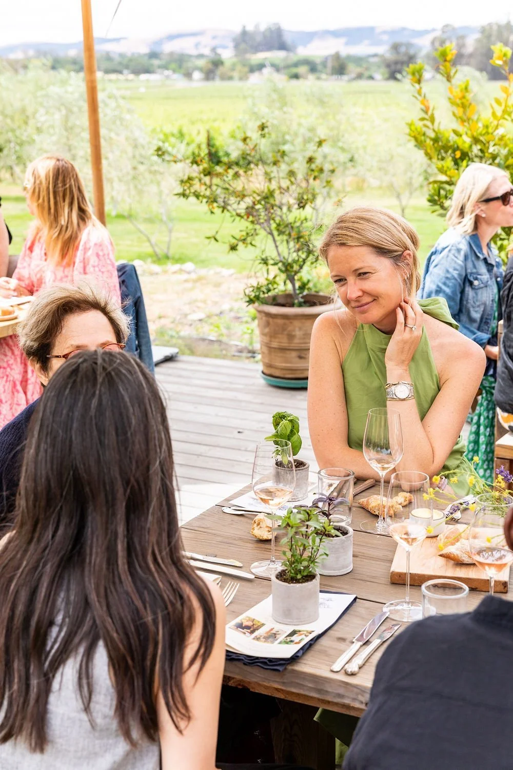 A group of women sitting at a table outdoors, engaging in conversation during a meal in a scenic garden or vineyard setting.