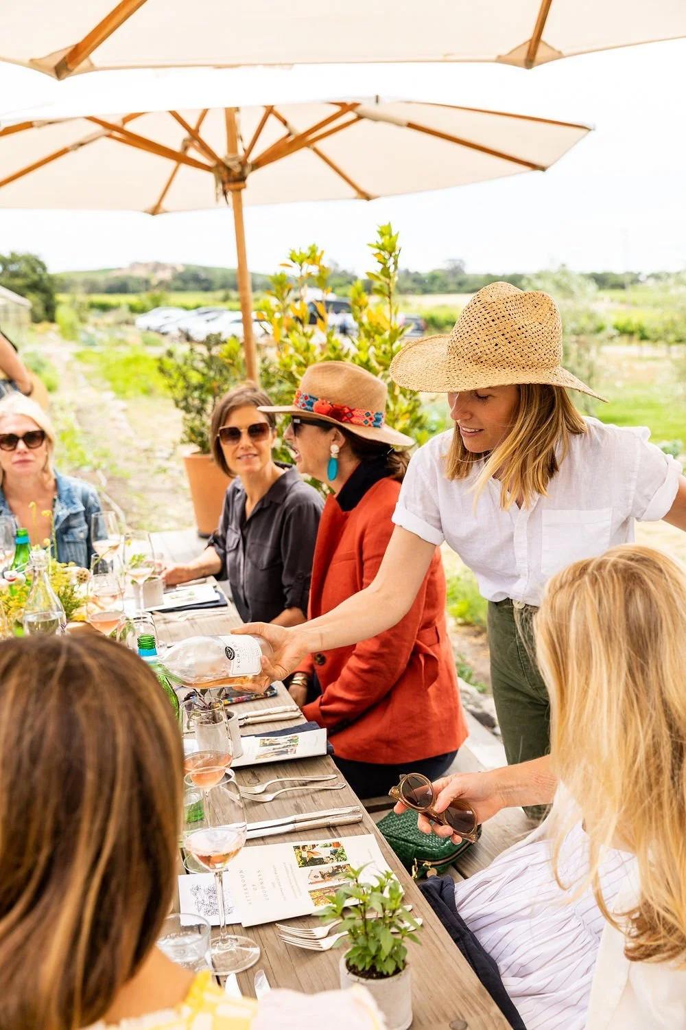 A group of women enjoying an outdoor meal at a vineyard, with one woman pouring water into a glass, under a large patio umbrella surrounded by greenery and a scenic landscape.