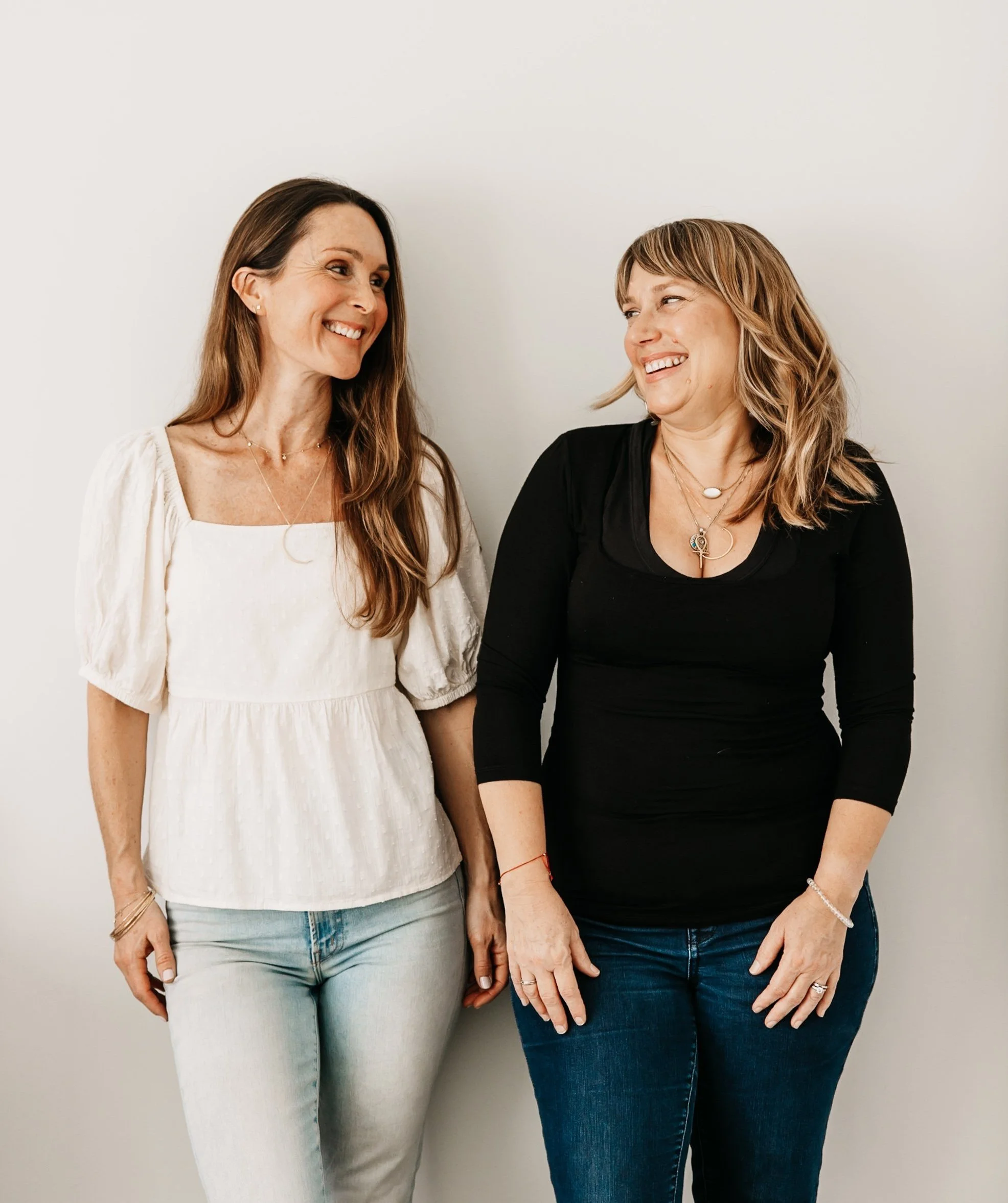 Two women standing together, smiling and looking at each other against a plain white wall.