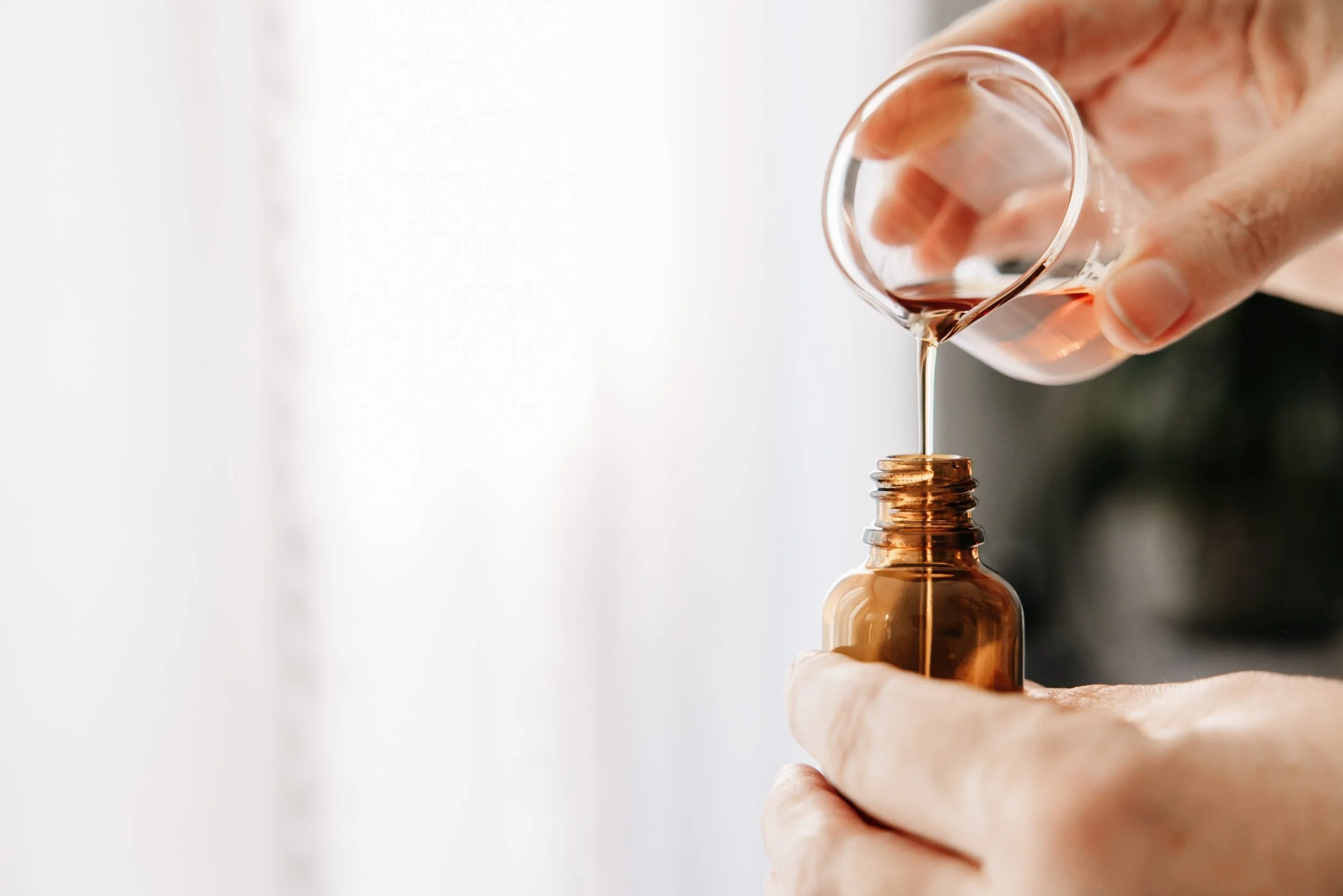 Person pouring pink liquid from a small glass container into a brown glass bottle.