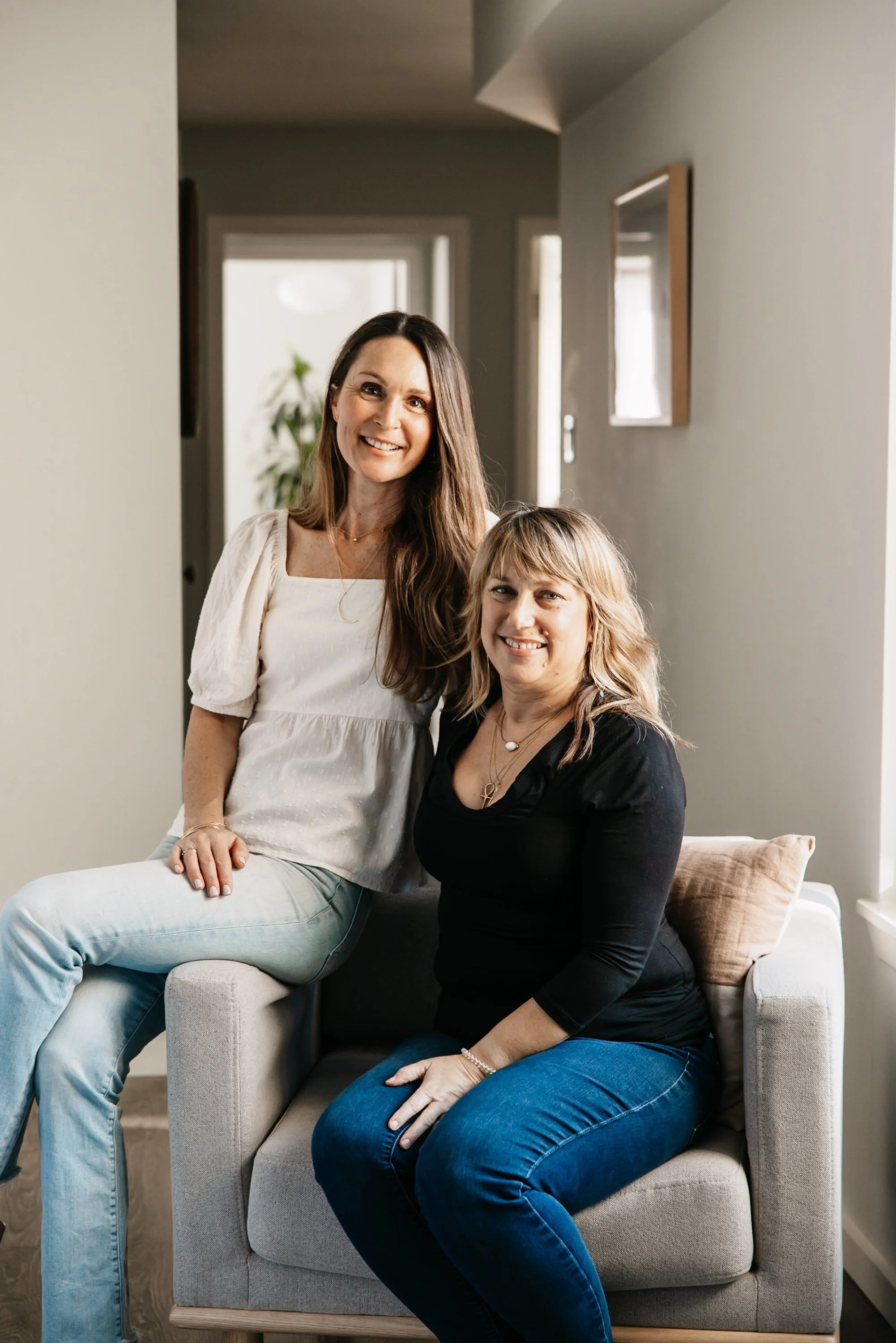 Two women smiling, one sitting on a beige armchair and the other sitting on the armrest, in a bright living room.
