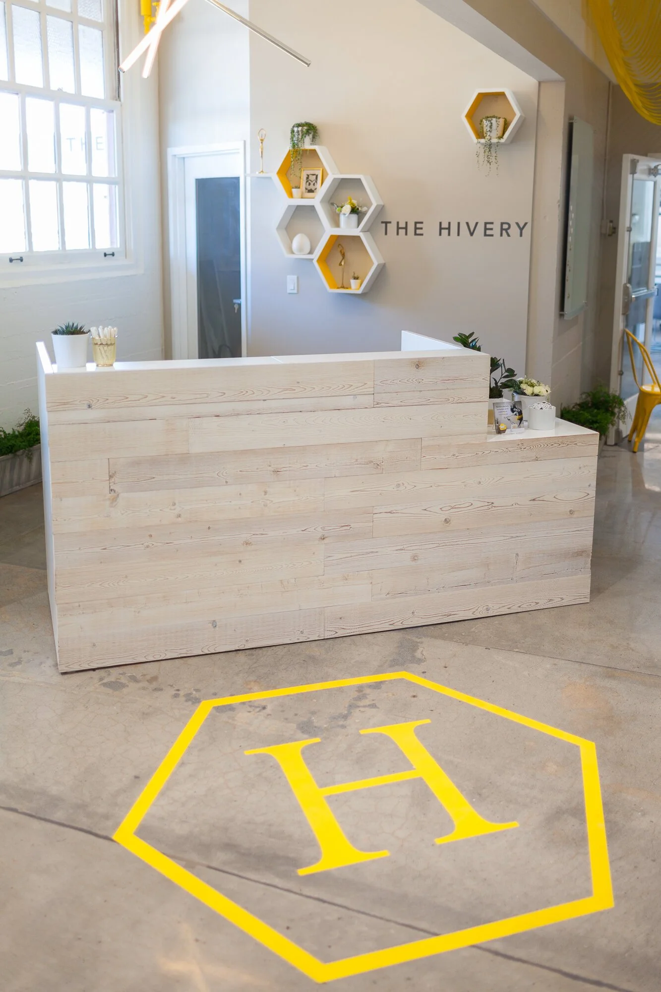 Modern reception desk made of light wood in a bright, welcoming space with decorative wall shelves and a yellow and white color scheme. There is a large yellow letter 'H' painted on the concrete floor.