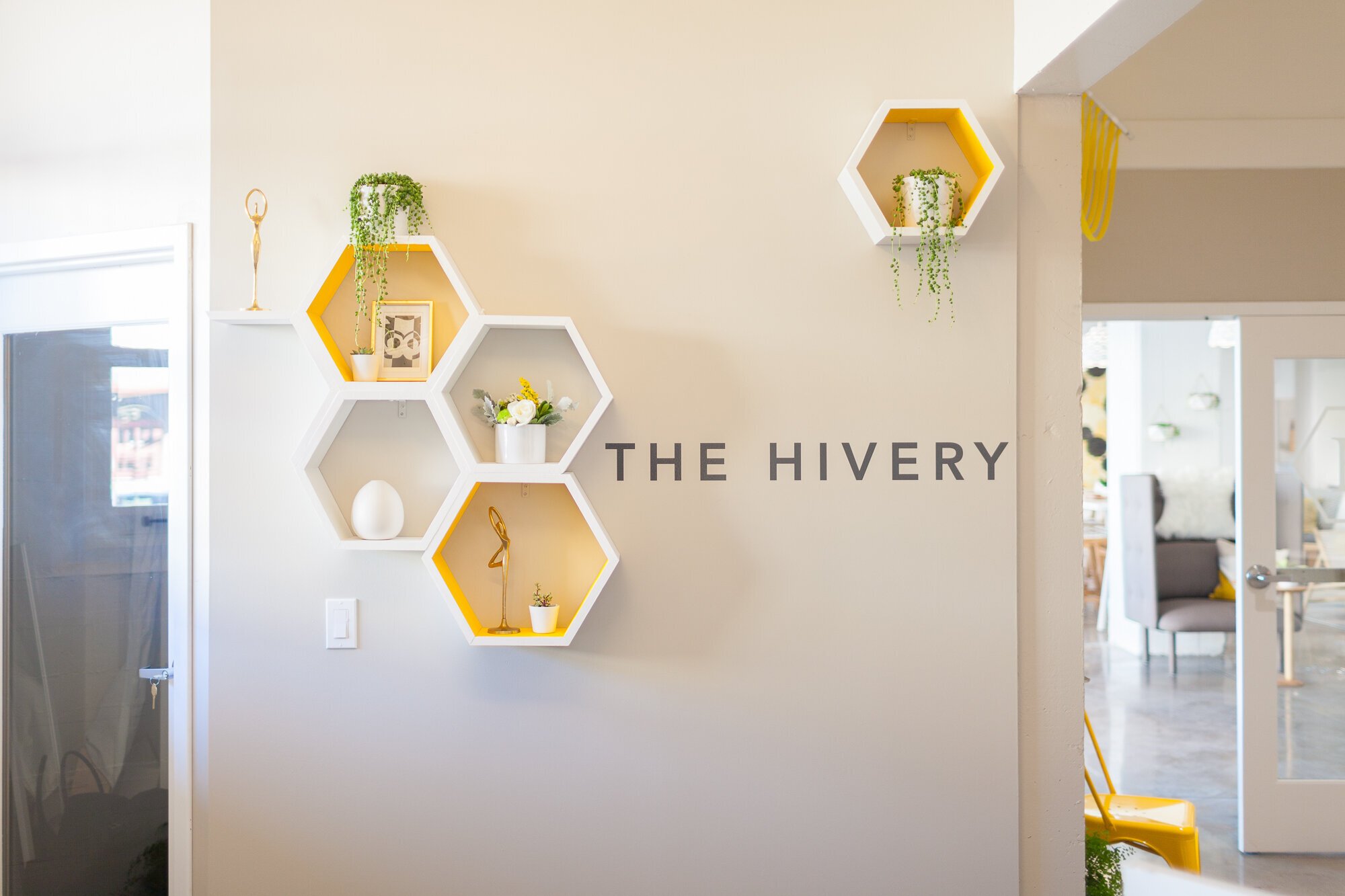 Wall with white and yellow hexagonal shelves decorated with plants and art, and black letters spelling 'THE HIVERY' on the wall.