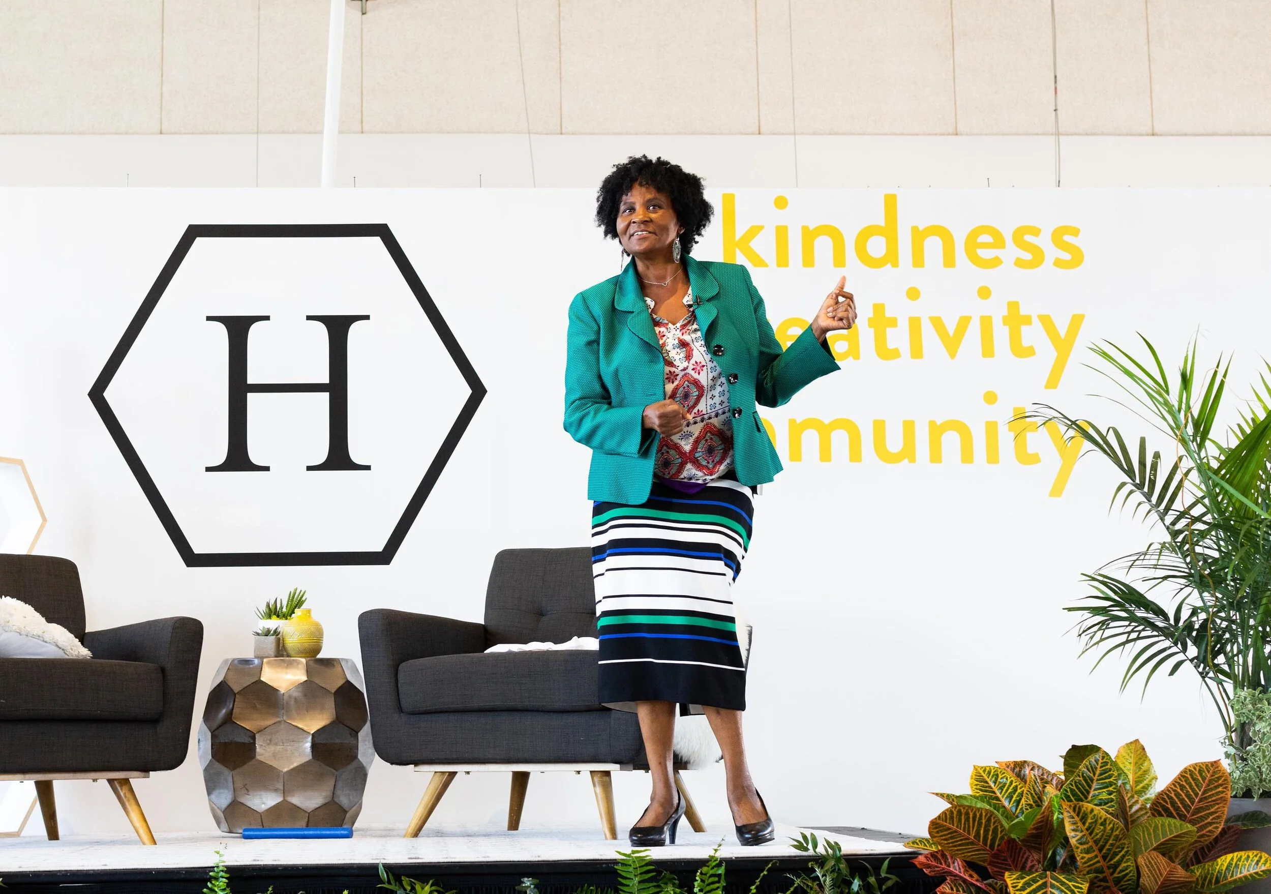 A woman standing on a stage with a backdrop that has large text reading 'kindness, creativity, community' and a logo with the letter H inside a hexagon. She is wearing a teal blazer, a patterned blouse, and a striped skirt, and she appears to be spea