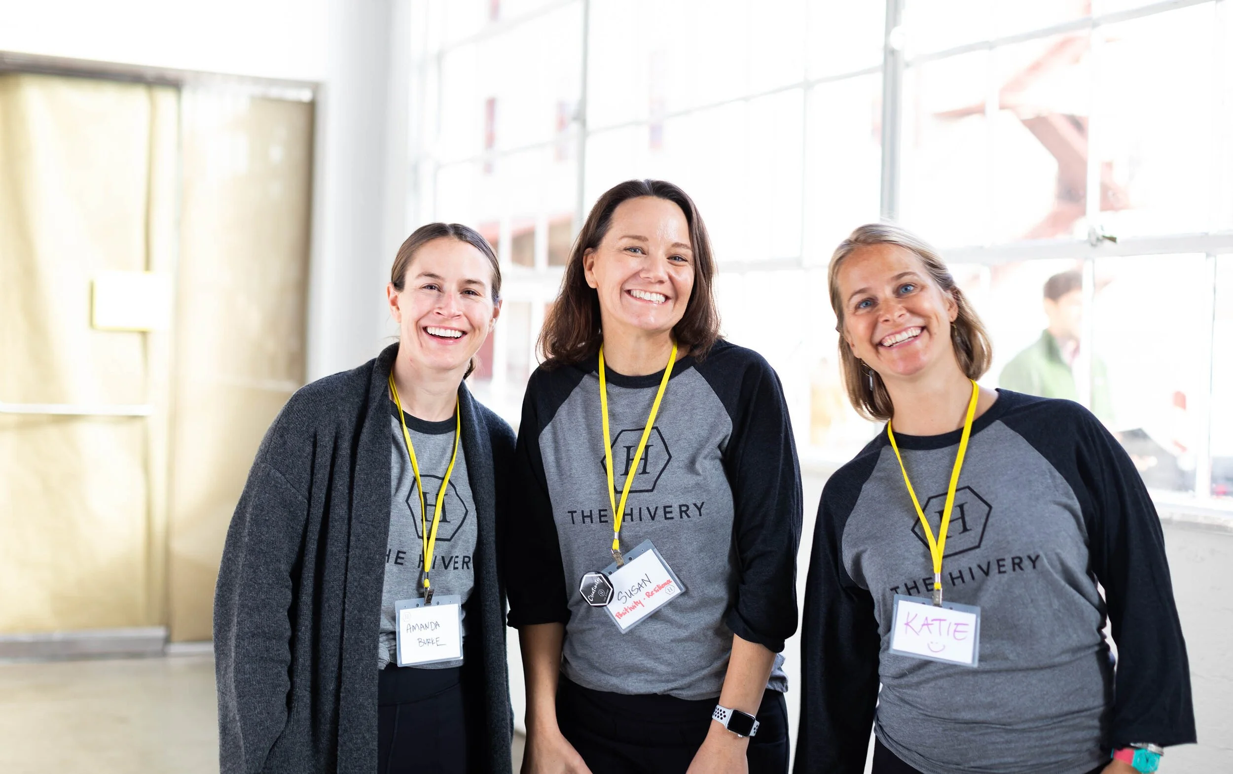 Three women smiling and wearing matching gray and black T-shirts with the words "The Thivery" and a hexagon logo. They are standing indoors in front of windows, with name tags on yellow lanyards around their necks.
