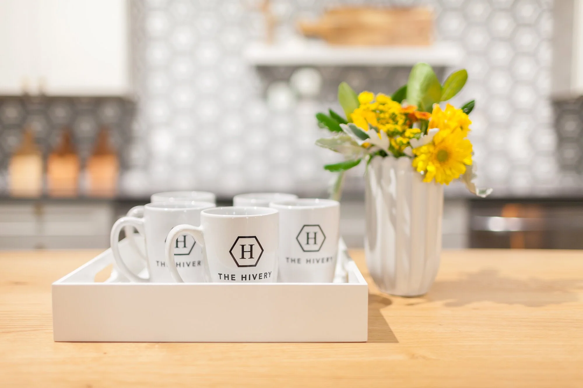 White mugs with the logo 'The Hivery' on a tray on a wooden table, with a vase of yellow and white flowers in the background.