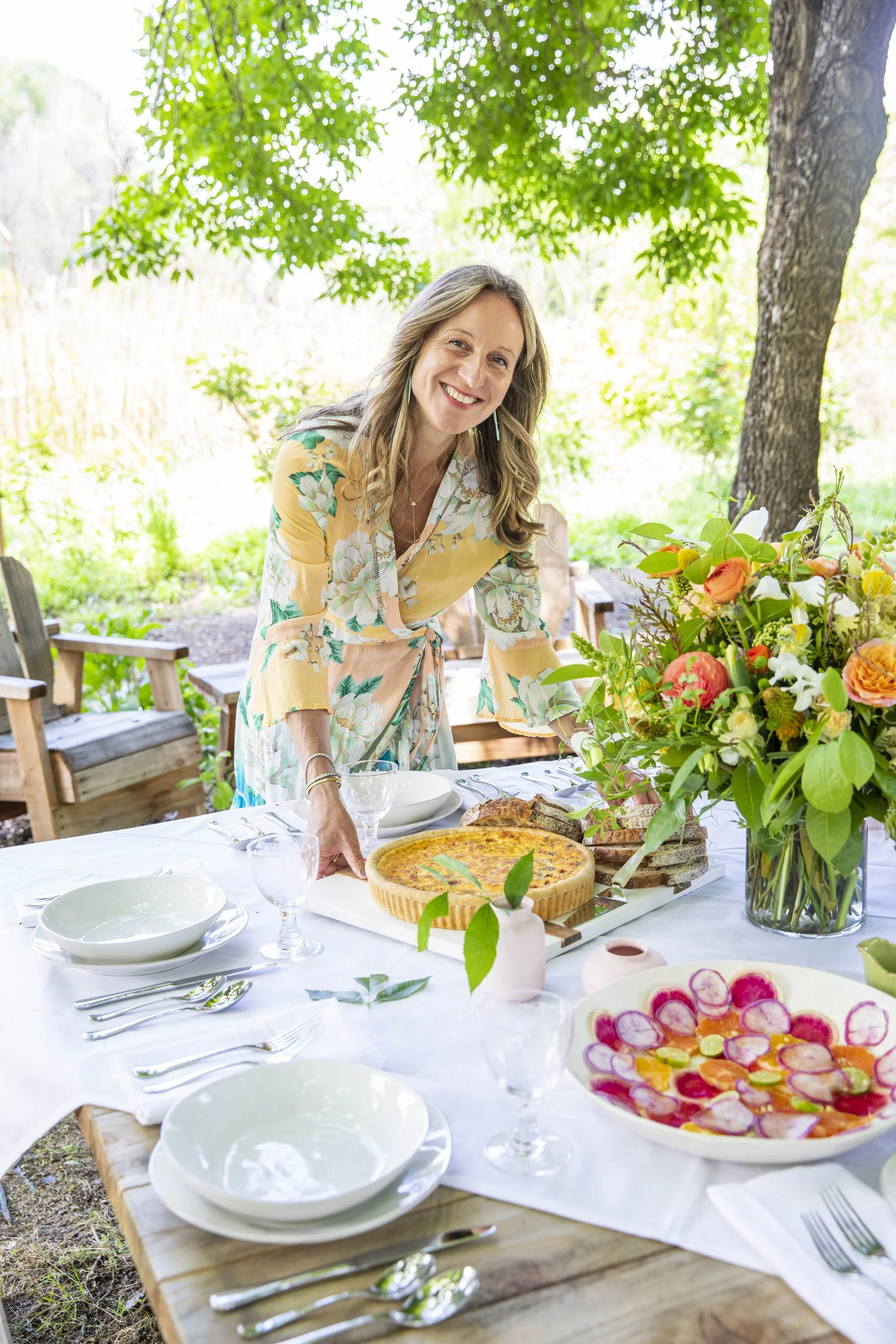 A woman in a yellow floral dress smiling and setting a table outdoors with a large bouquet of colorful flowers, a pie, and a salad.