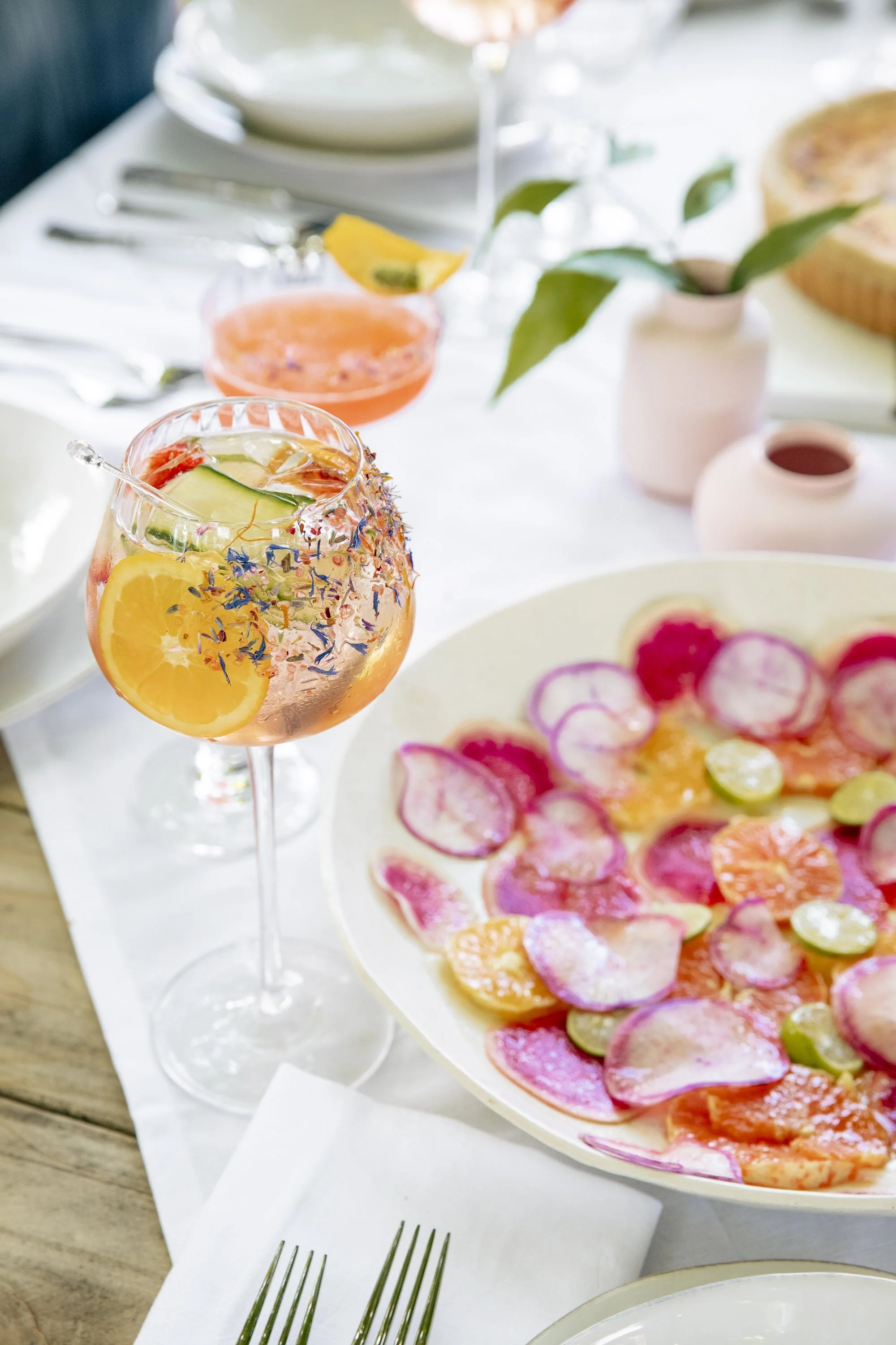 A table set for brunch with colorful drinks, a floral salad, a plant, and tableware on a white tablecloth.