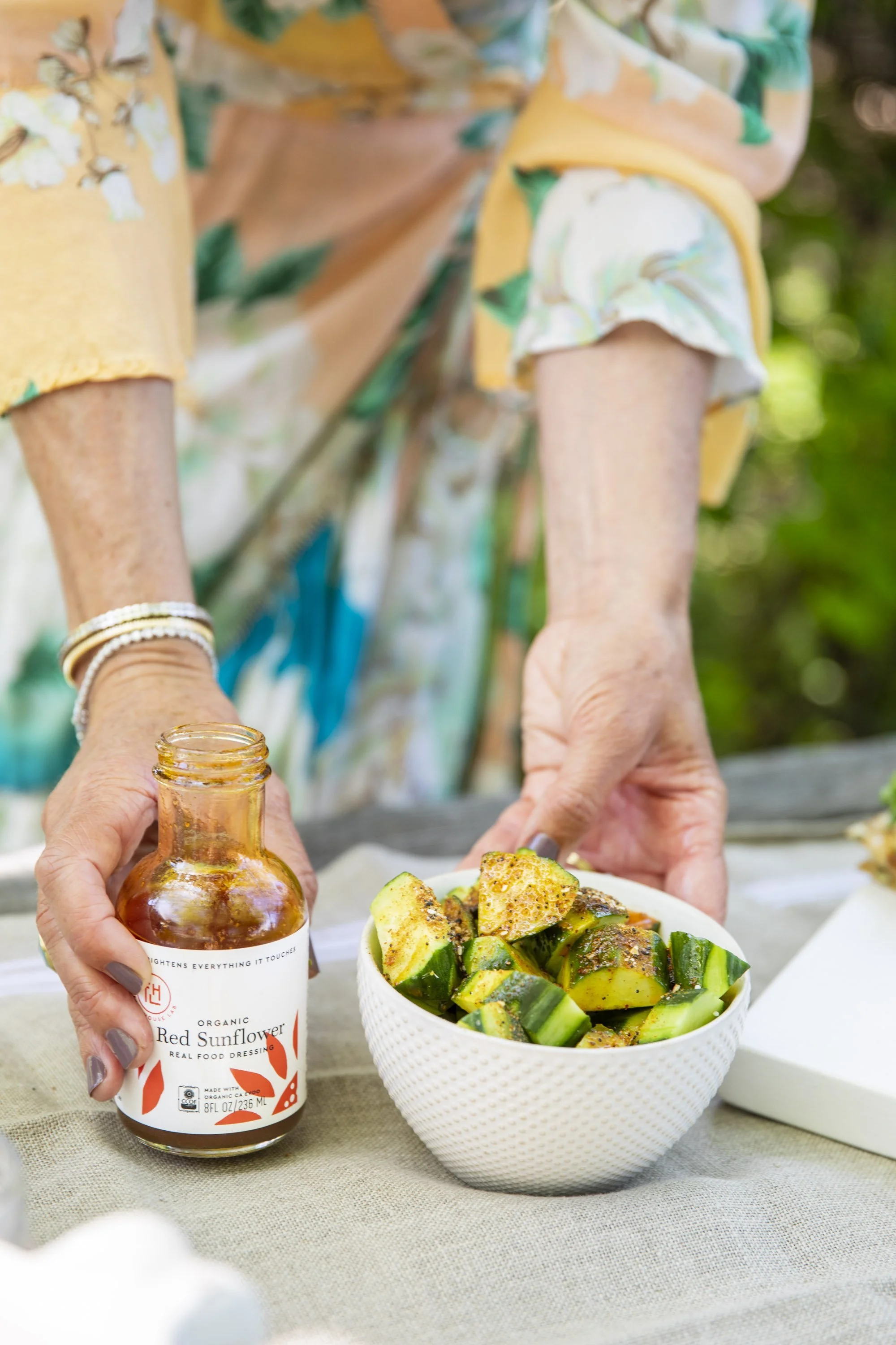 A person in floral clothing arranges a bowl of cucumber salad on a table, with a bottle of red sunflower organic salad dressing nearby.