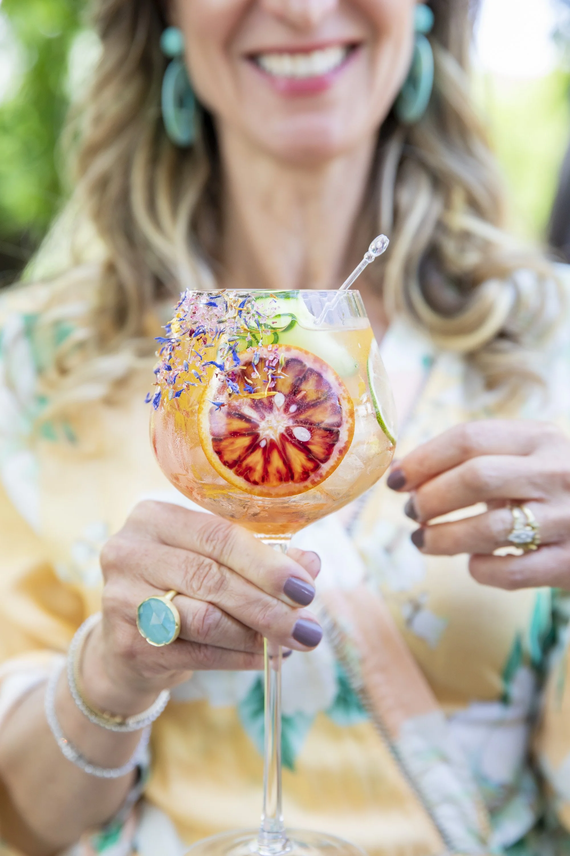 A smiling woman holding a large glass of colorful cocktail with slices of blood orange, lime, and cucumber, garnished with edible flowers.