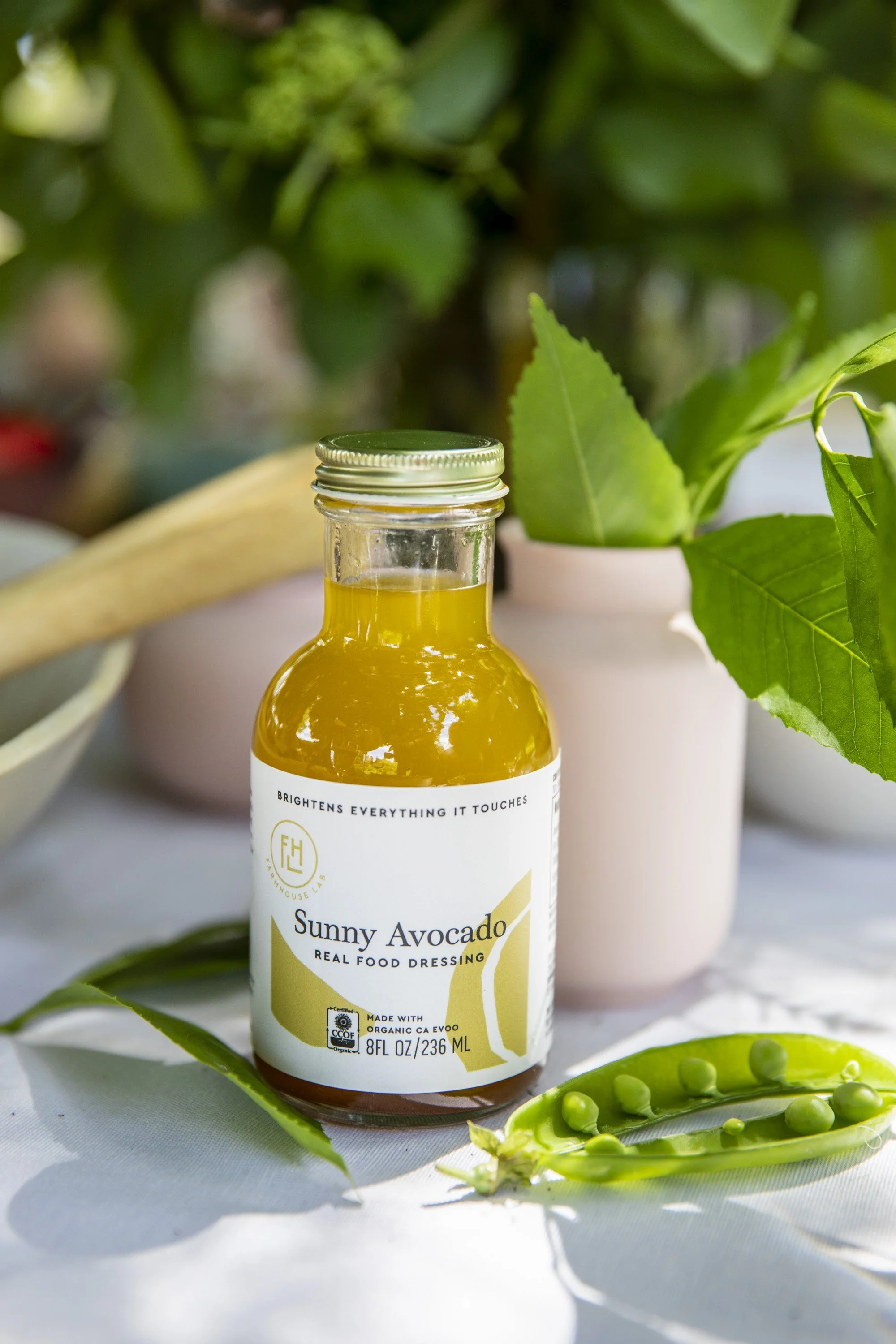 Bottle of Sunny Avocado dressing with green leaves and pea pod on a white surface.