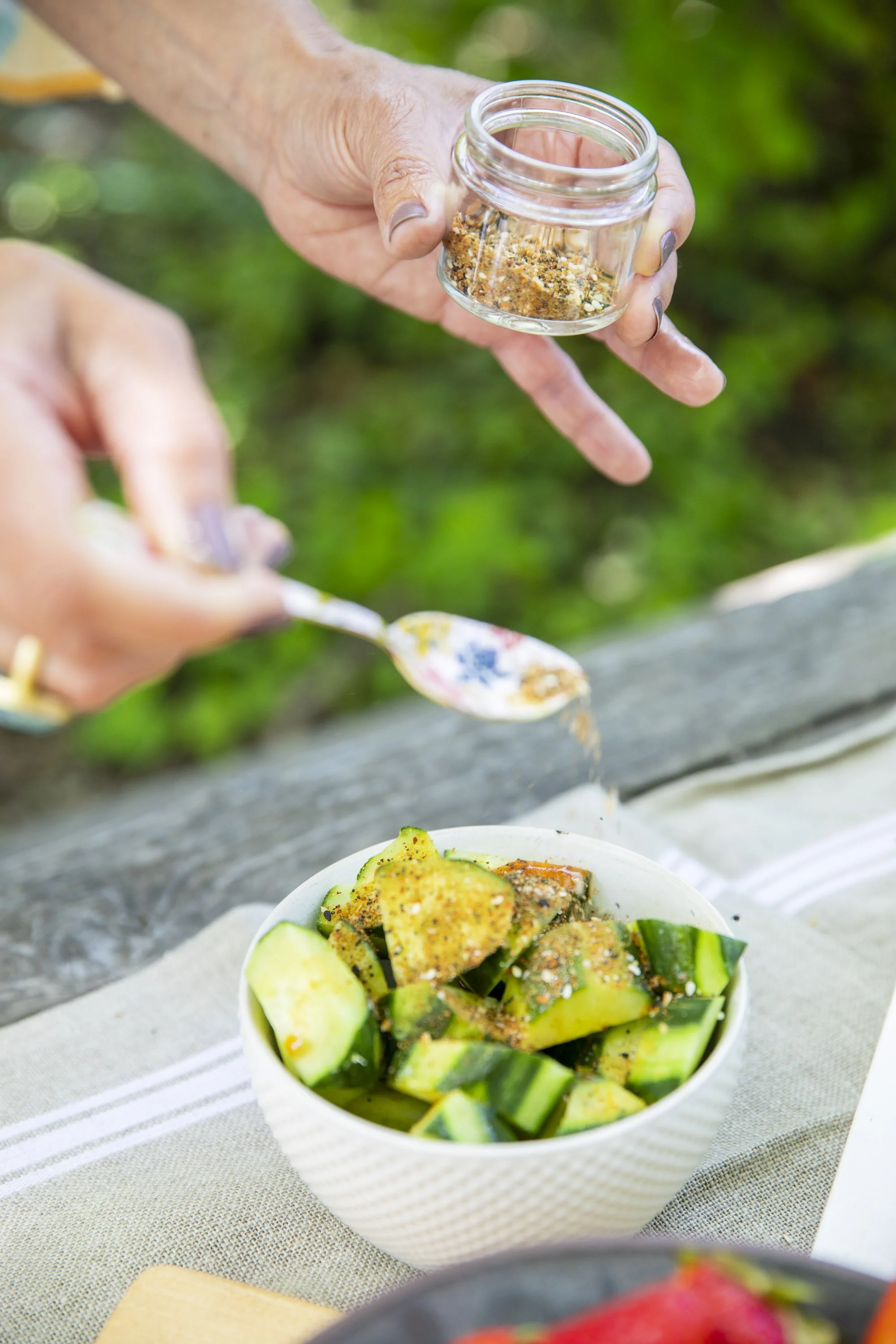 Person sprinkling seasoning on a bowl of sliced cucumbers and other vegetables outdoors.