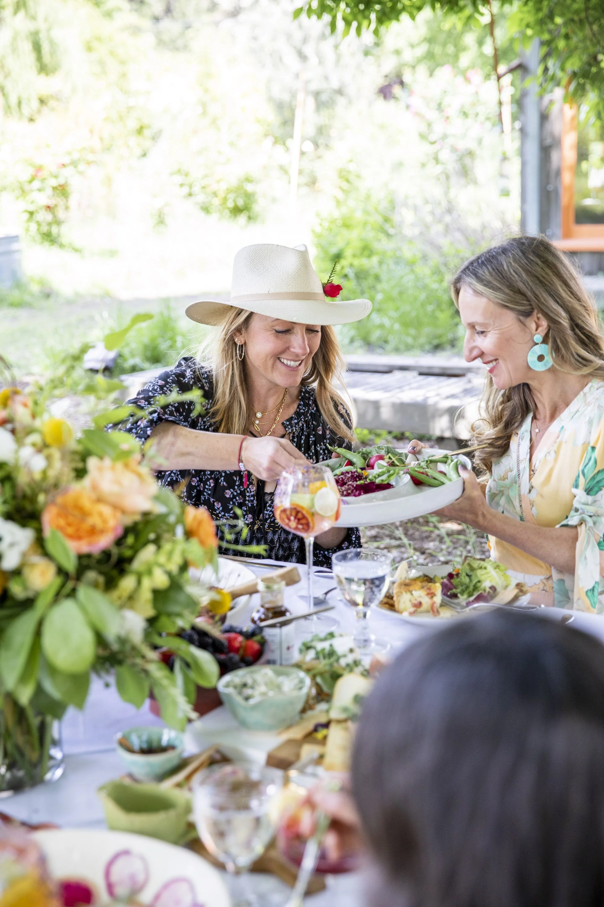 Two women smiling and sharing food at an outdoor garden party, surrounded by flowers, with a table filled with food and drinks.
