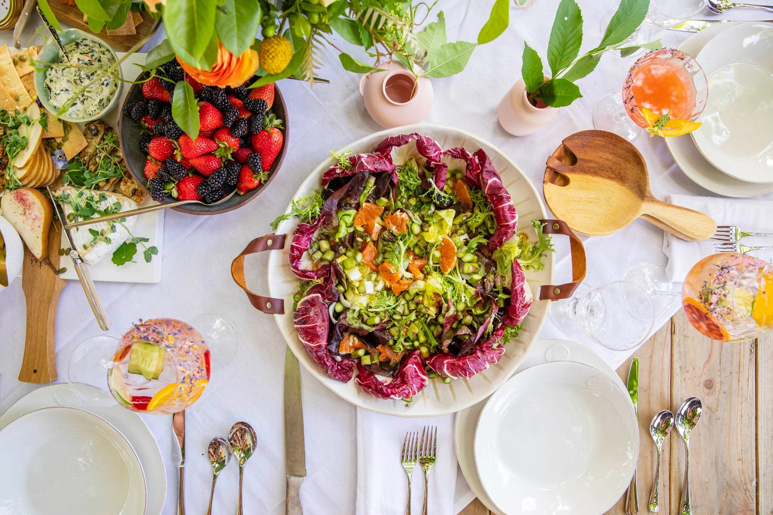 A top-down view of a dining table set with various dishes, including a large mixed green salad in the center, bowls of berries and cheese, and colorful drinks.