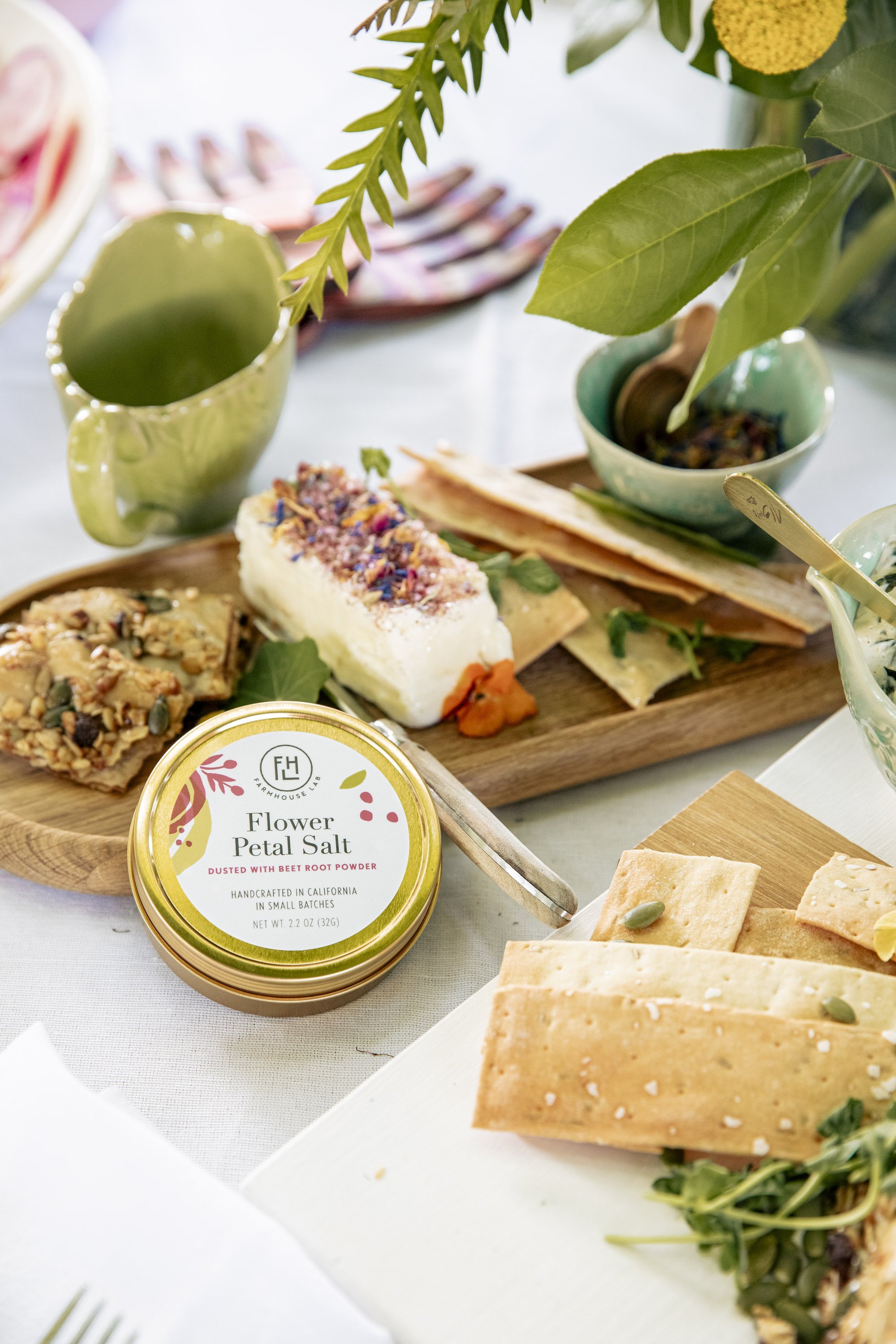 A table with a variety of foods, including crackers, cheese, a small bowl of nuts, a container of flower petal salt, and a cheese block topped with colorful edible petals, along with green leaves and a wooden serving board.