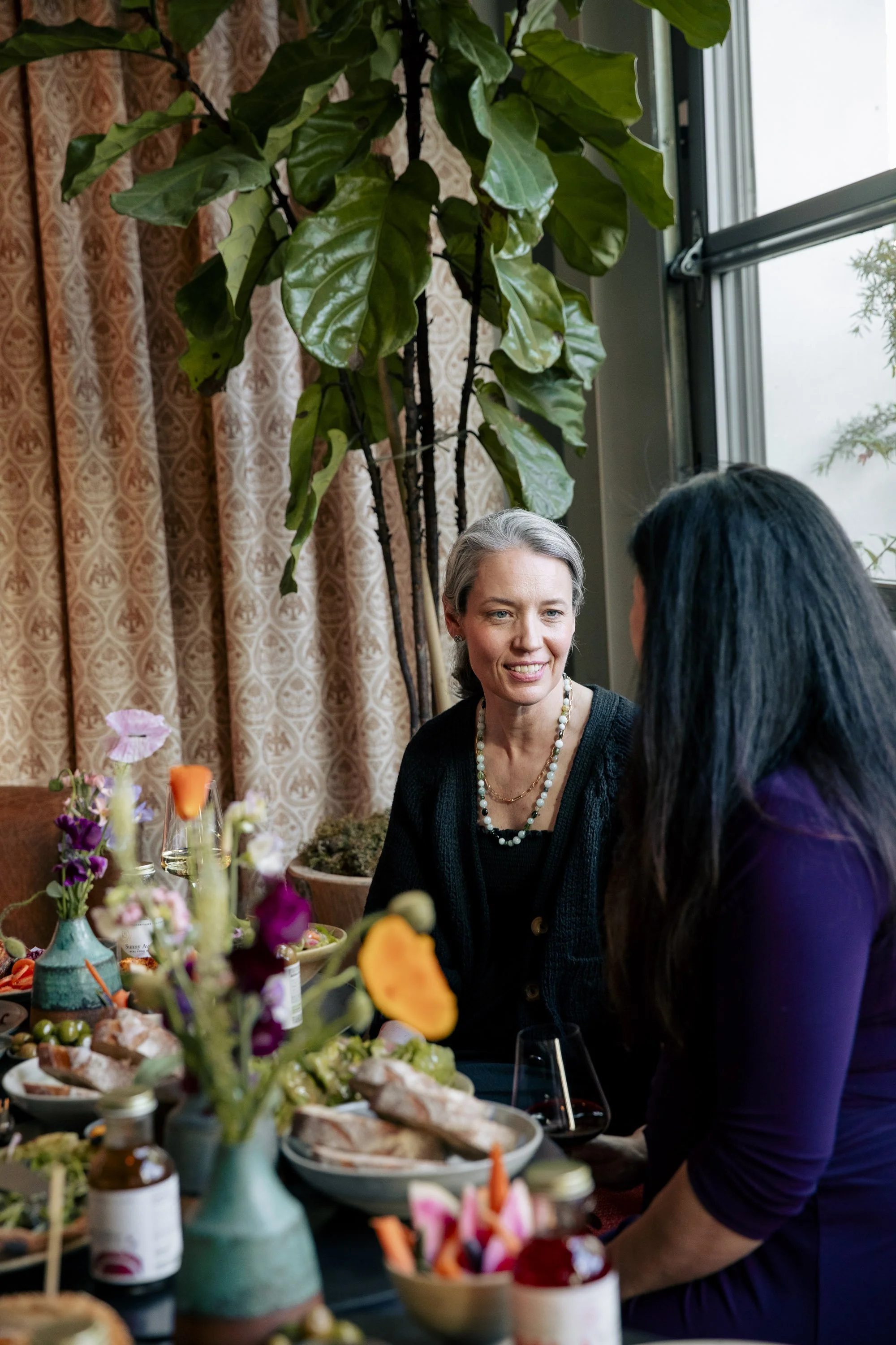 Two women having a conversation at a table with food and flowers, indoors near a large window and a tall houseplant.