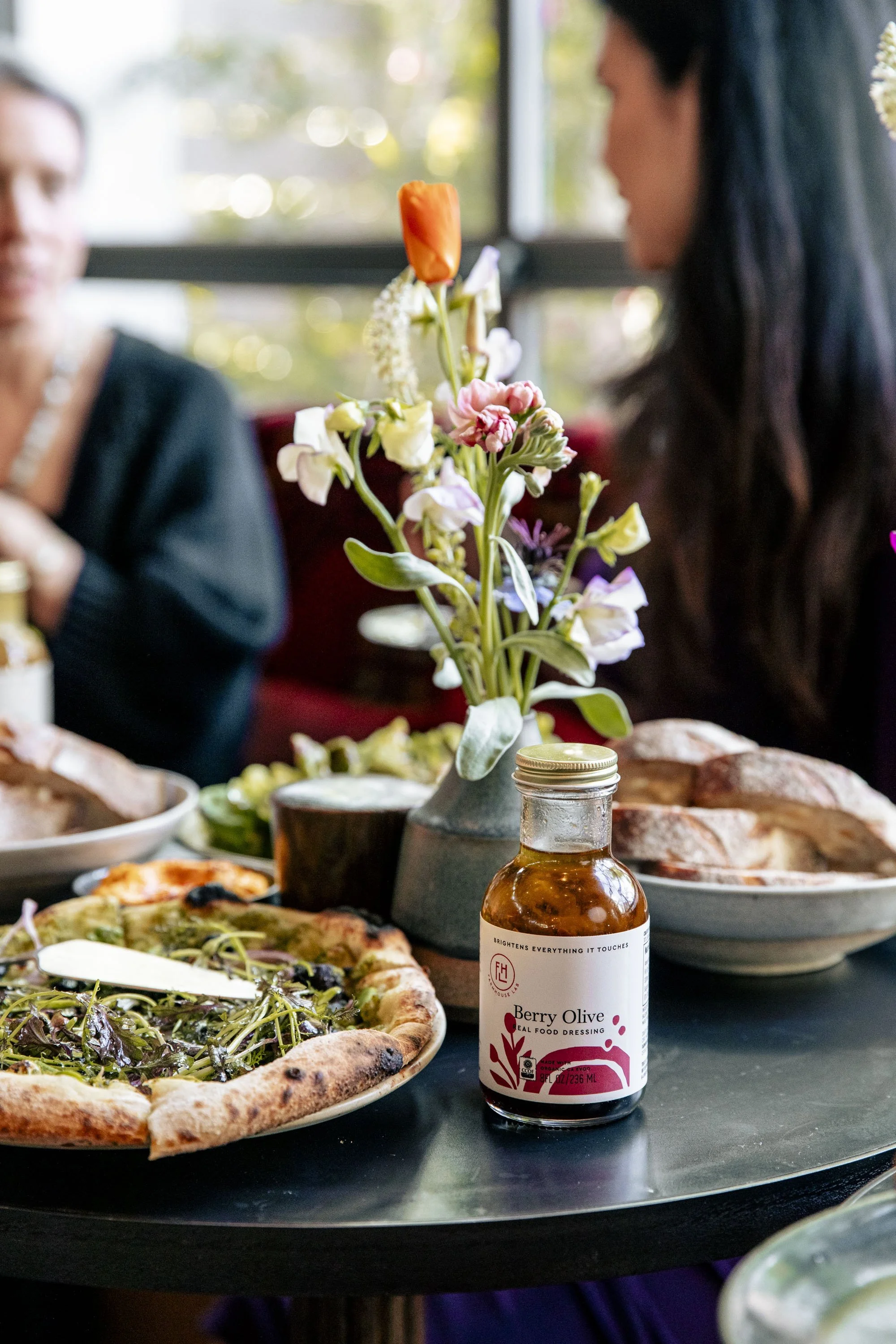 A table with a vase of colorful flowers, a bottle of berry olive salad dressing, pizza with greens, and bread, with two women seated in the background near a window.