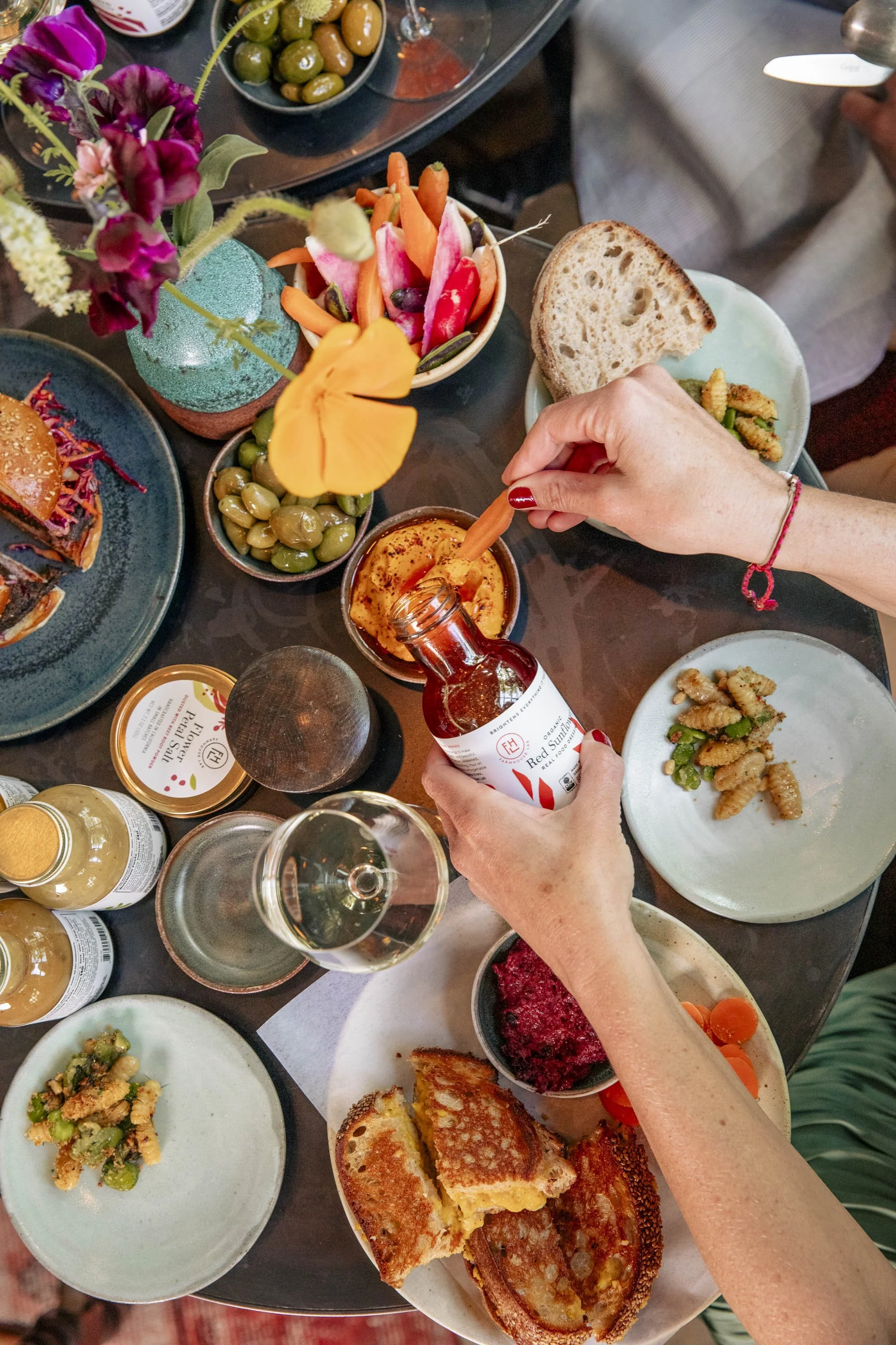 A table filled with various dishes including bread, olives, a bowl of salad, appetizers, a bottle of hot sauce, and a glass of white wine, with a person pouring hot sauce onto food.