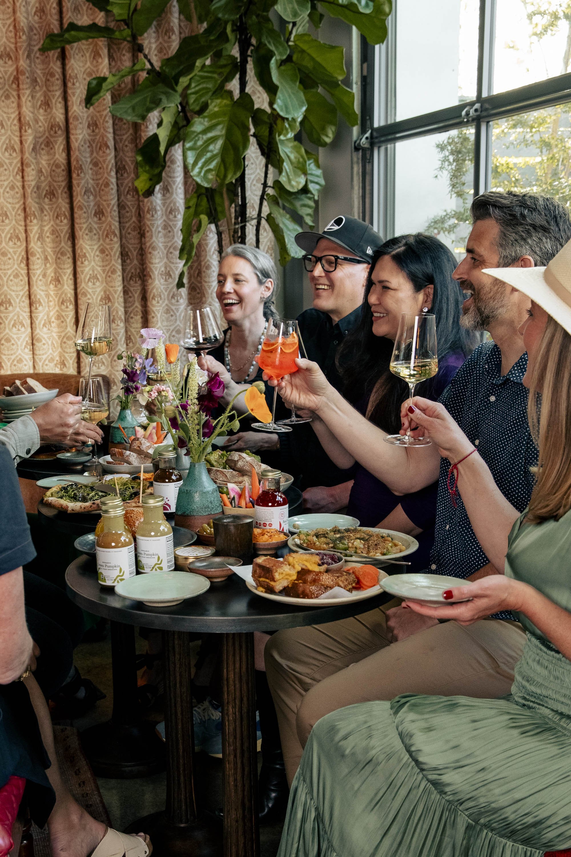 Group of people enjoying a dinner party with food and drinks at a decorated table in a well-lit room with large windows.