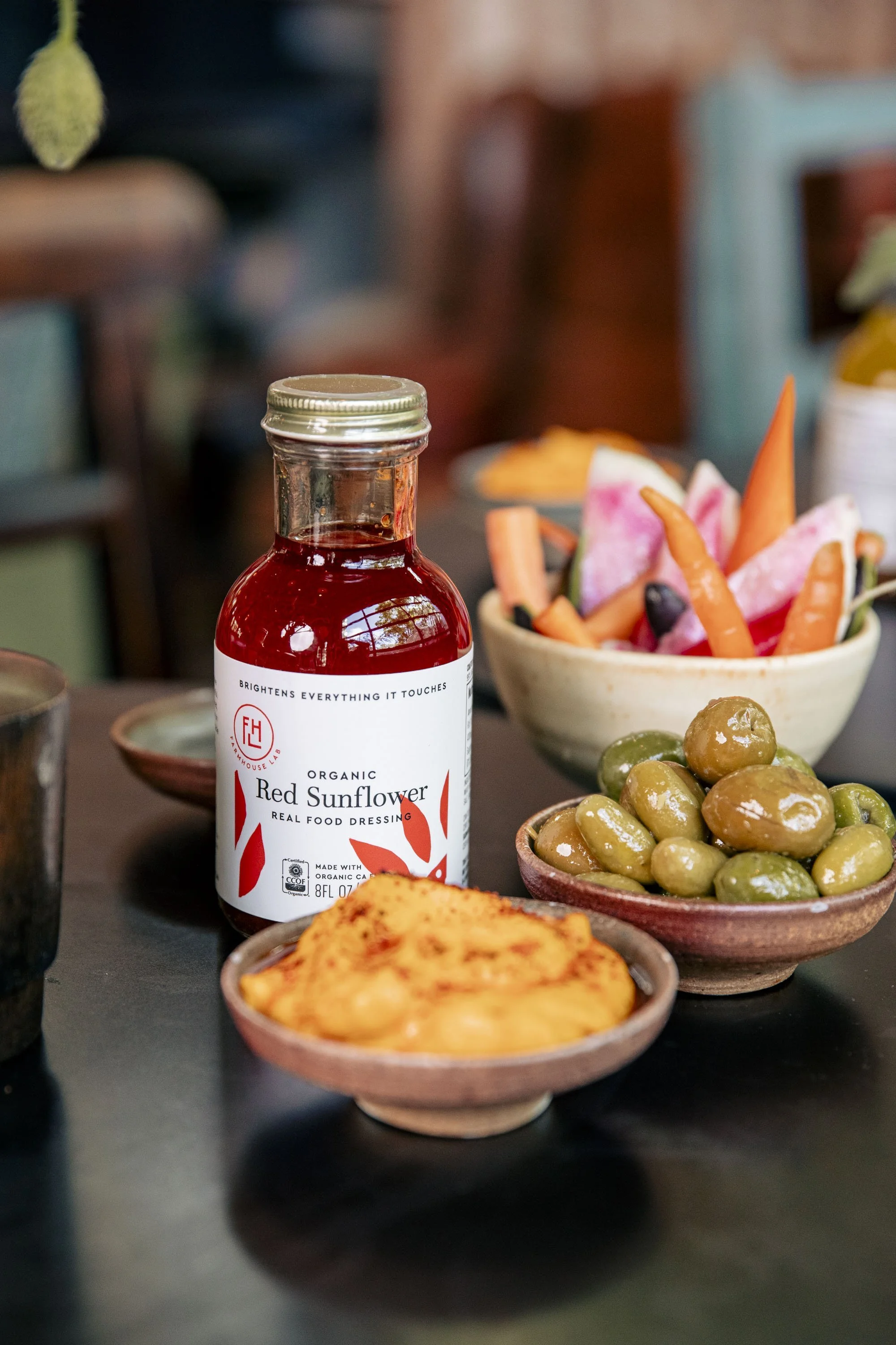 A table with a bottle of organic Red Sunflower dressing, a bowl of mixed olives, a bowl of colorful vegetable sticks, and a dish of mashed potatoes.