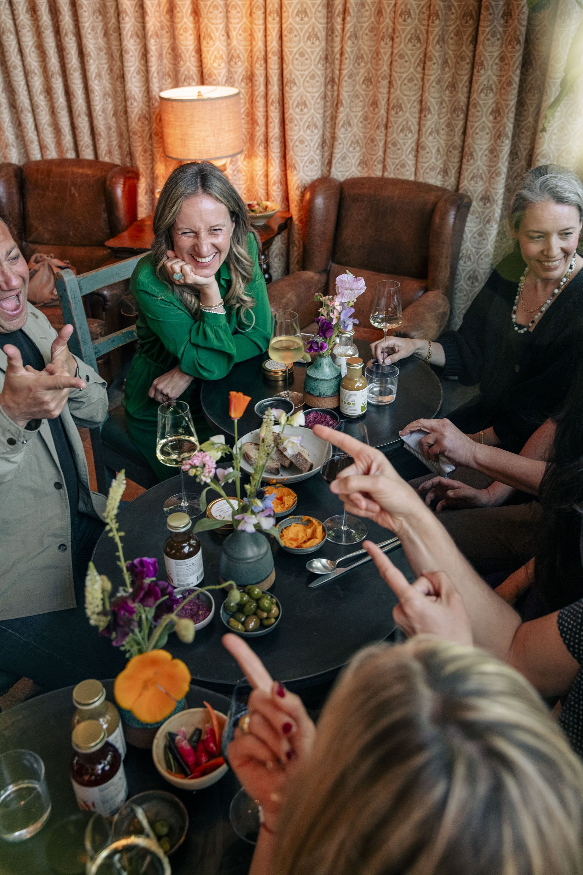 Group of people gathered around a table enjoying food and drinks, smiling and laughing, in a cozy room with curtains and a lamp.