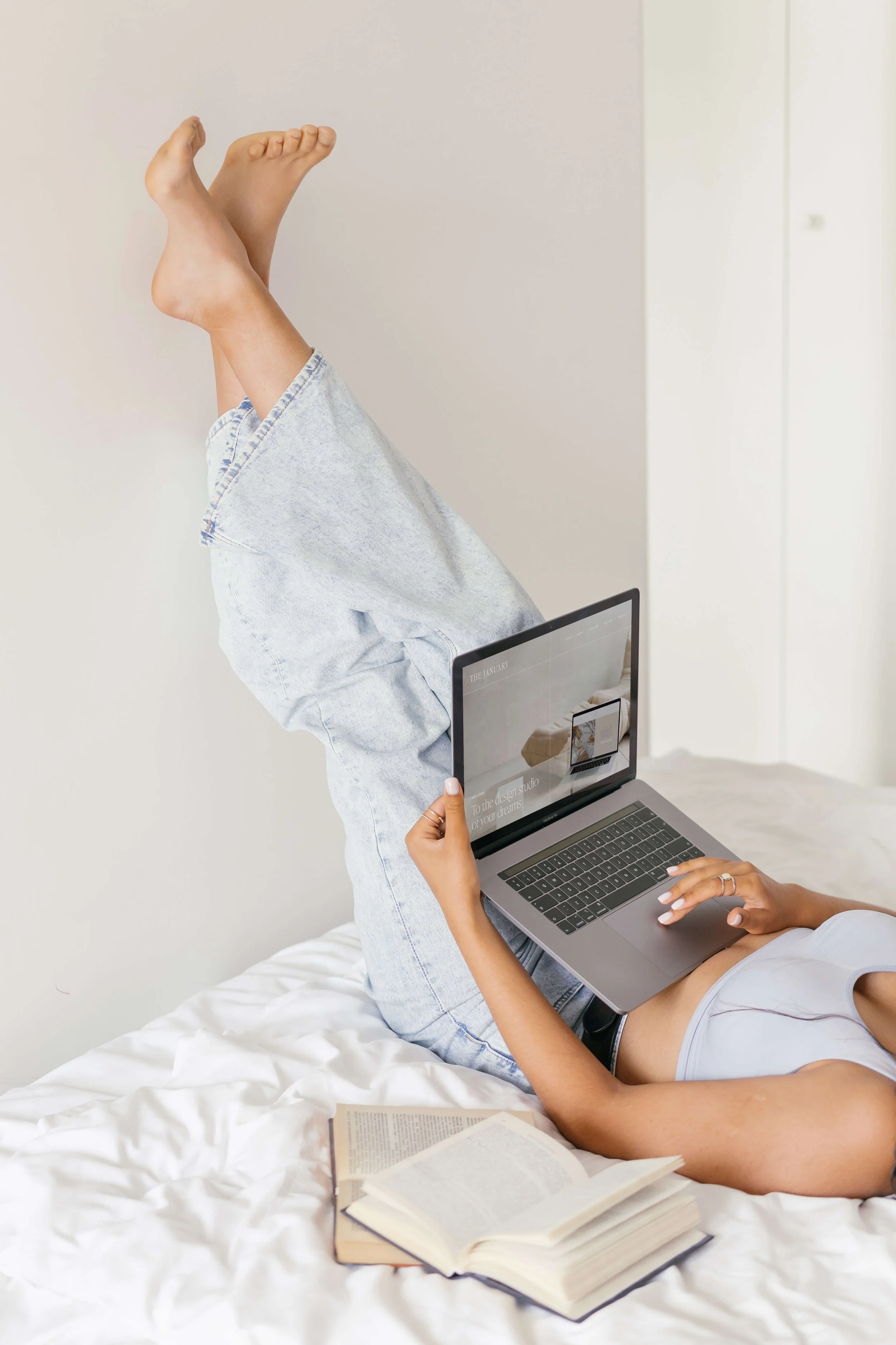 Person lying on bed with legs raised, using a laptop, with open books nearby, in a bright room.