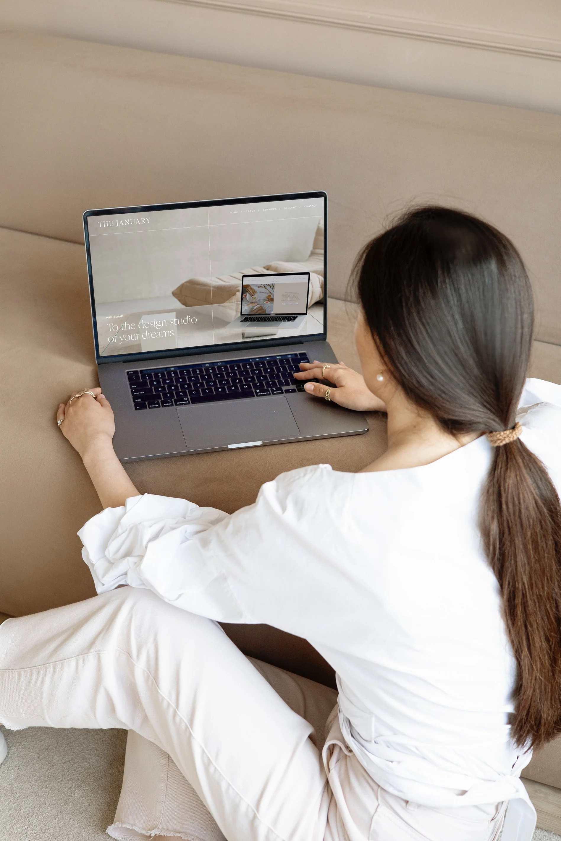 A woman with long brown hair, wearing a white top and beige pants, sitting on a beige couch and working on a silver laptop with a website featuring a beige bed and white text on the screen.