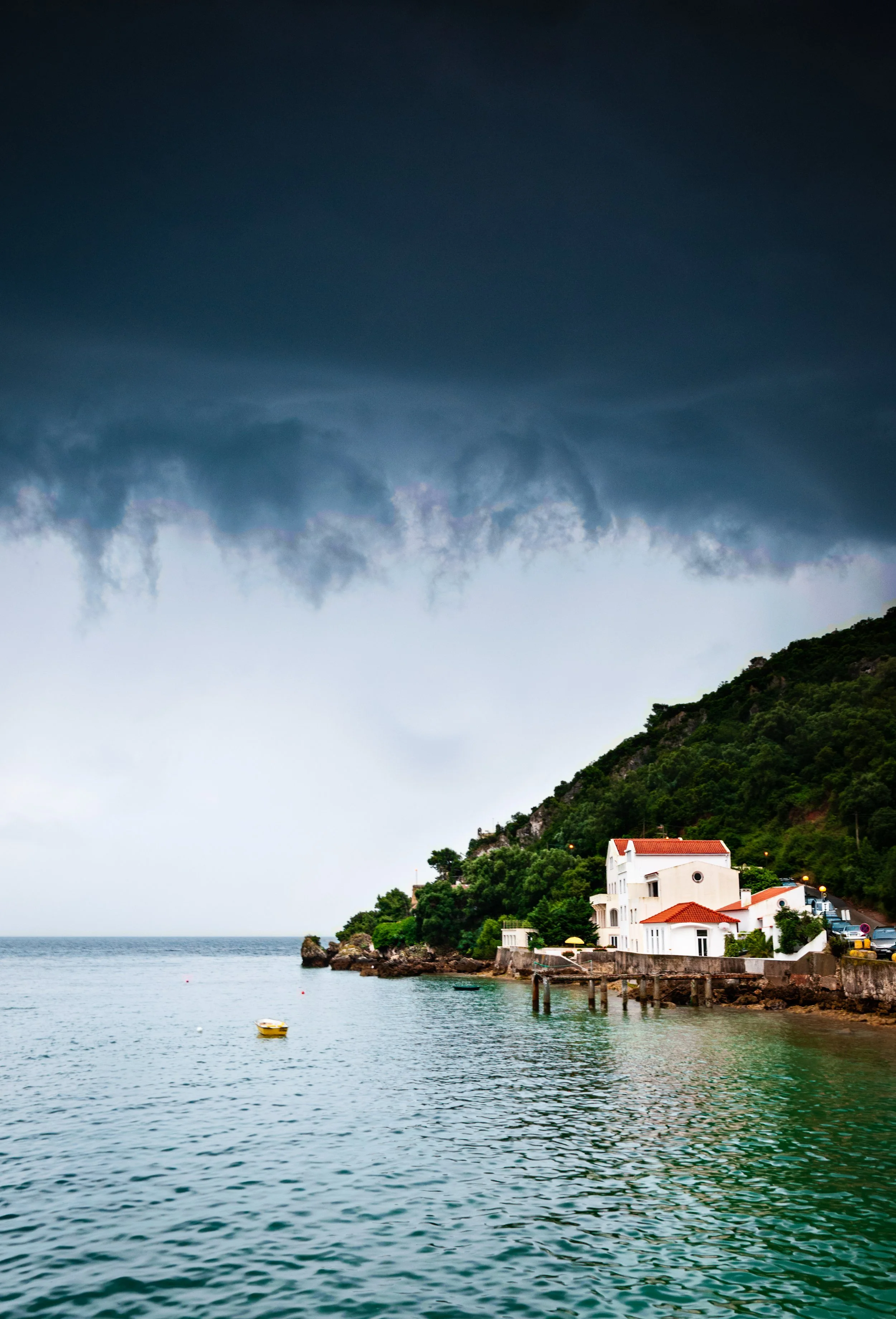 Storm clouds over a small coastal village with white buildings and red roofs, surrounded by greenery, near calm water with a small yellow boat.