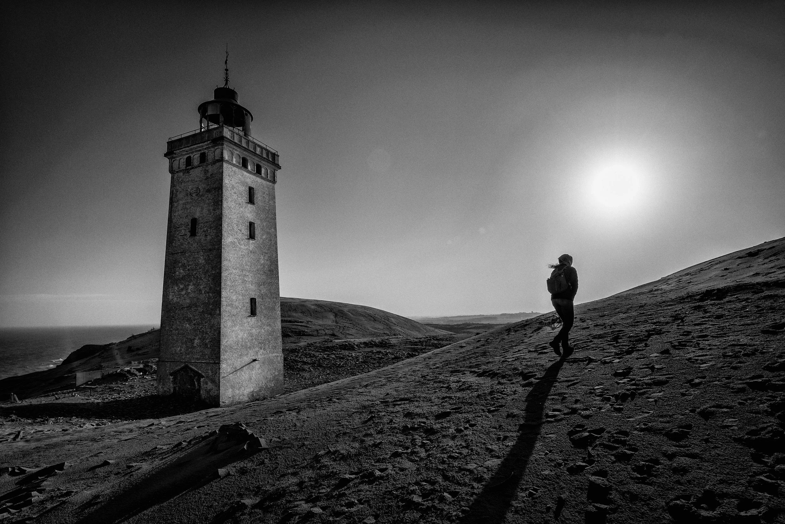 A person walking on a sloped sandy terrain near a lighthouse at sunset or sunrise, in black and white.