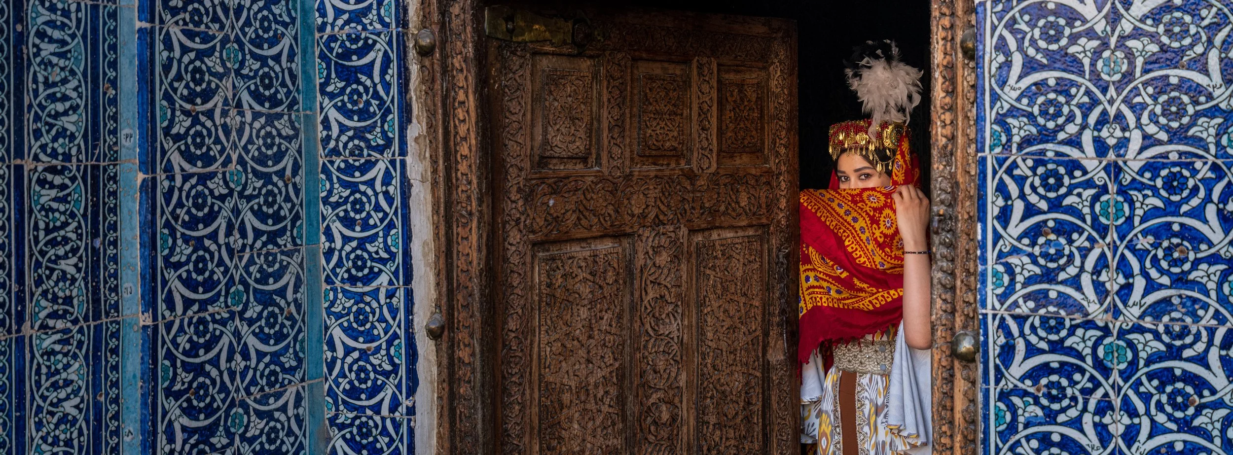 A woman dressed in traditional Southeast Asian attire, wearing a red and yellow scarf and headpiece with white feathers, peeks out from behind an intricately carved wooden door framed by blue and white patterned tile walls.