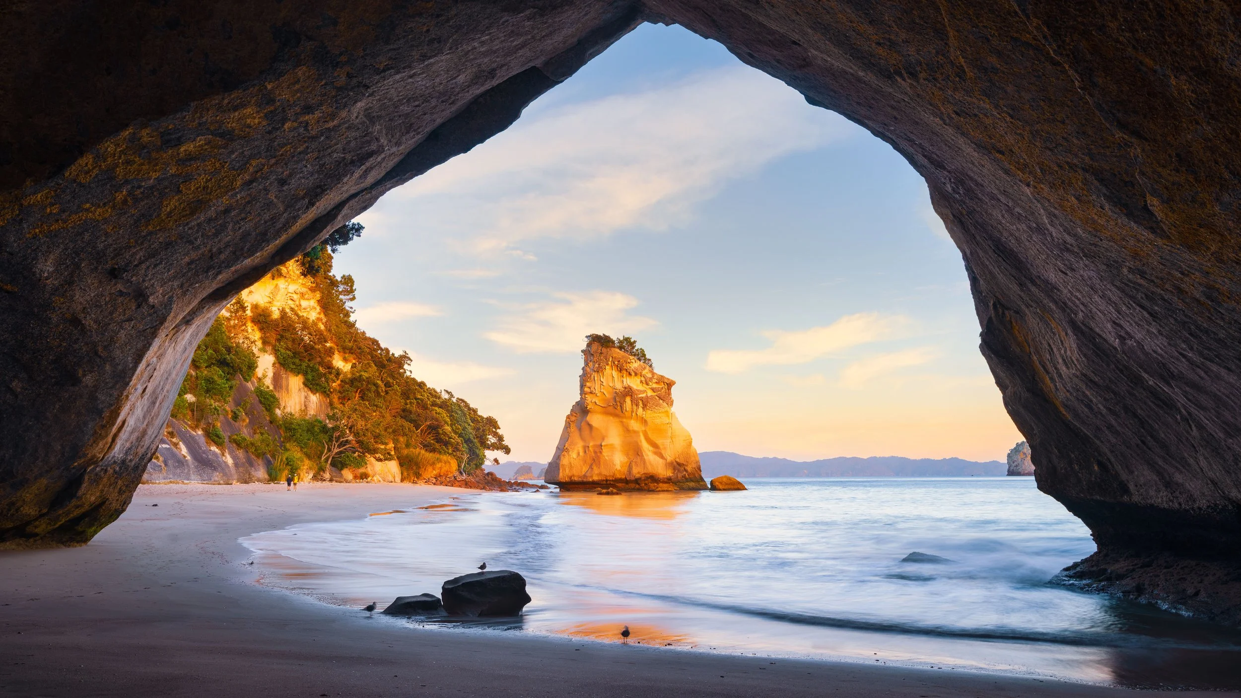 View of a beach with a large rock formation in the water, seen through a cave opening, with trees on the cliff and a partly cloudy sky.