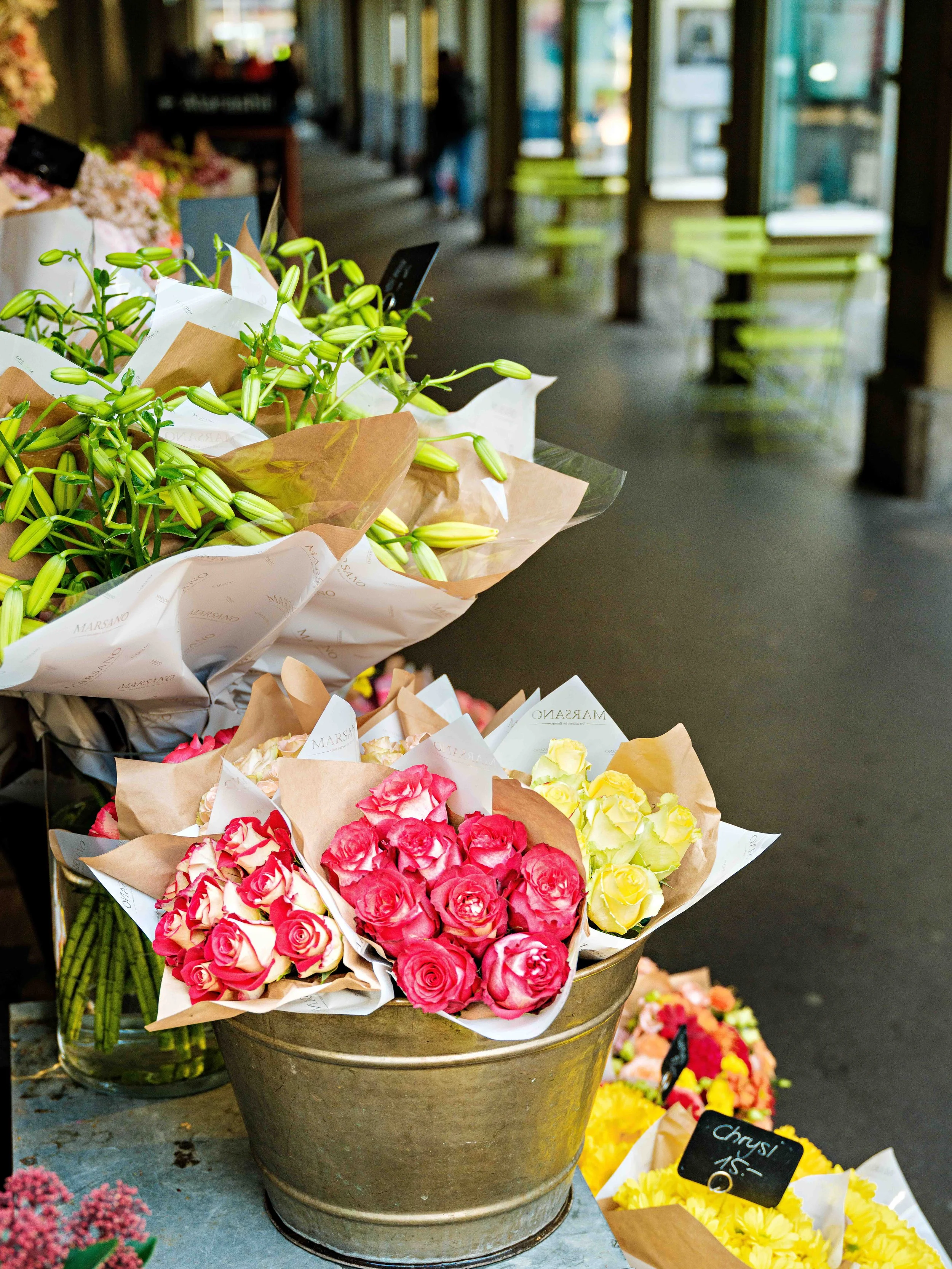 Vases and buckets of pink, yellow, and white flowers outside a flower shop, with a sidewalk and store interior in the background.