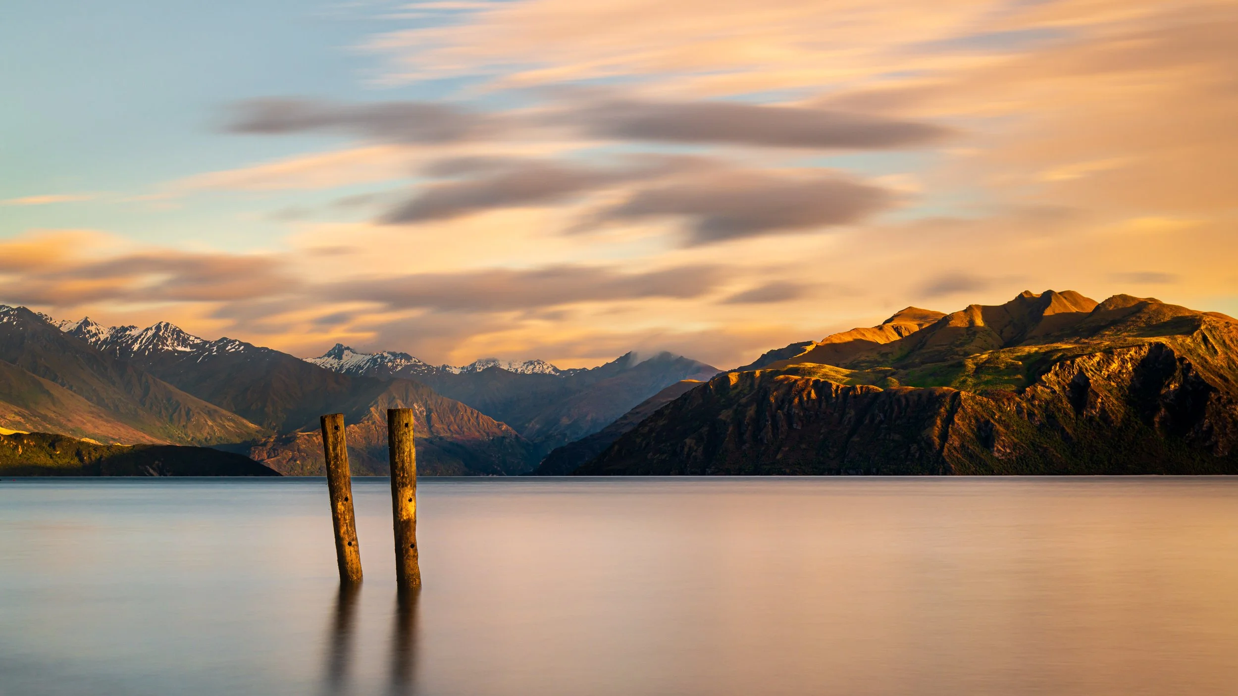 A serene lake at sunset with snow-capped mountains in the background and two wooden posts emerging from calm water.
