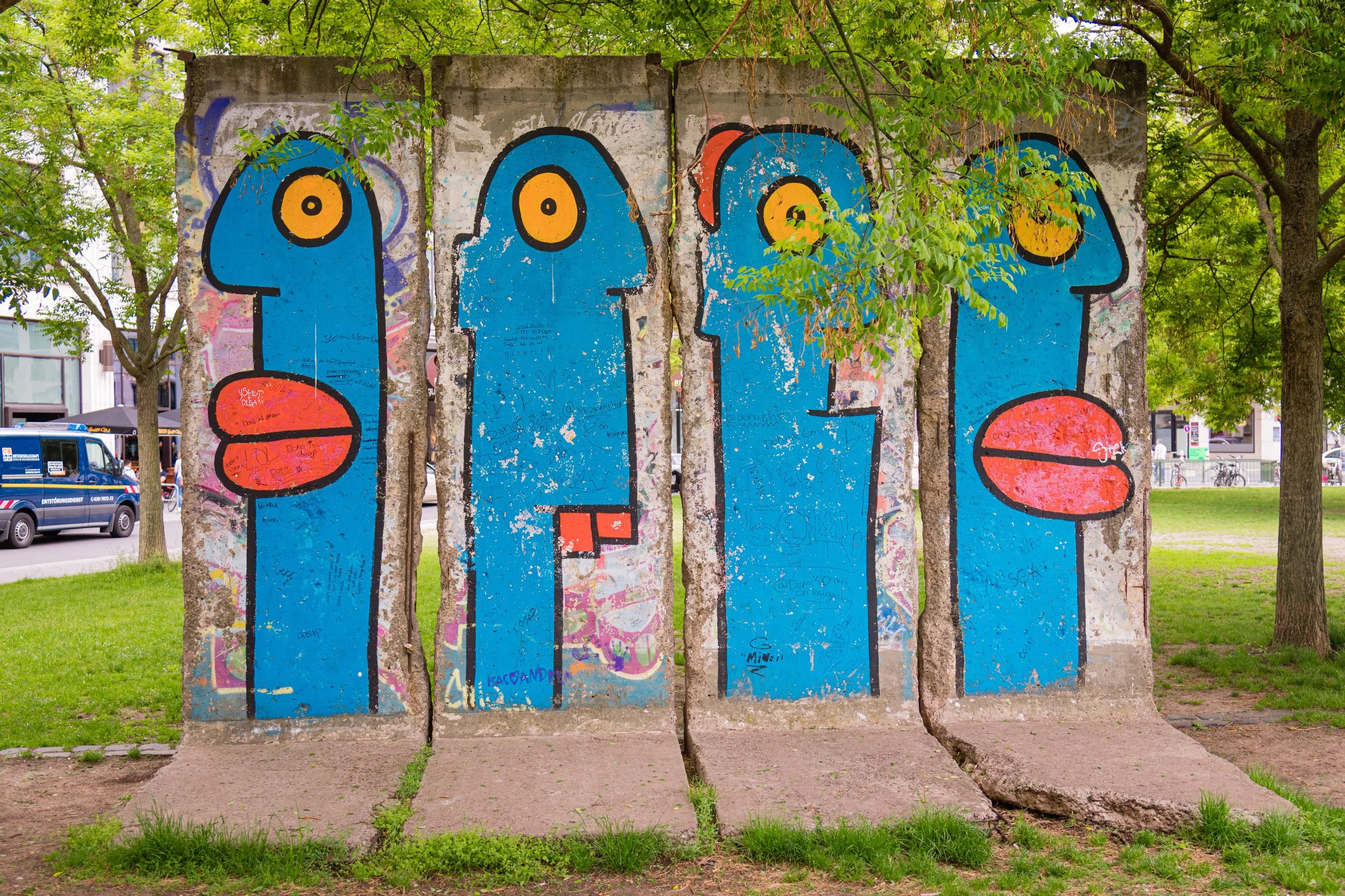 Street art graffiti of four blue abstract faces with yellow eyes and red lips on a concrete wall, surrounded by green trees in an outdoor park.