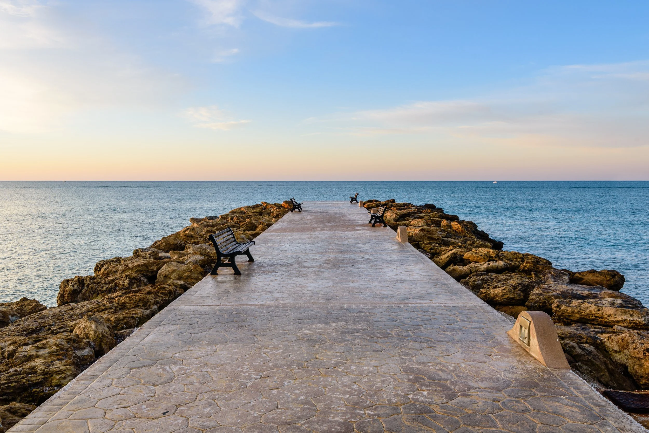 A stone pier with benches extending into a calm ocean, under a clear sky with soft clouds.