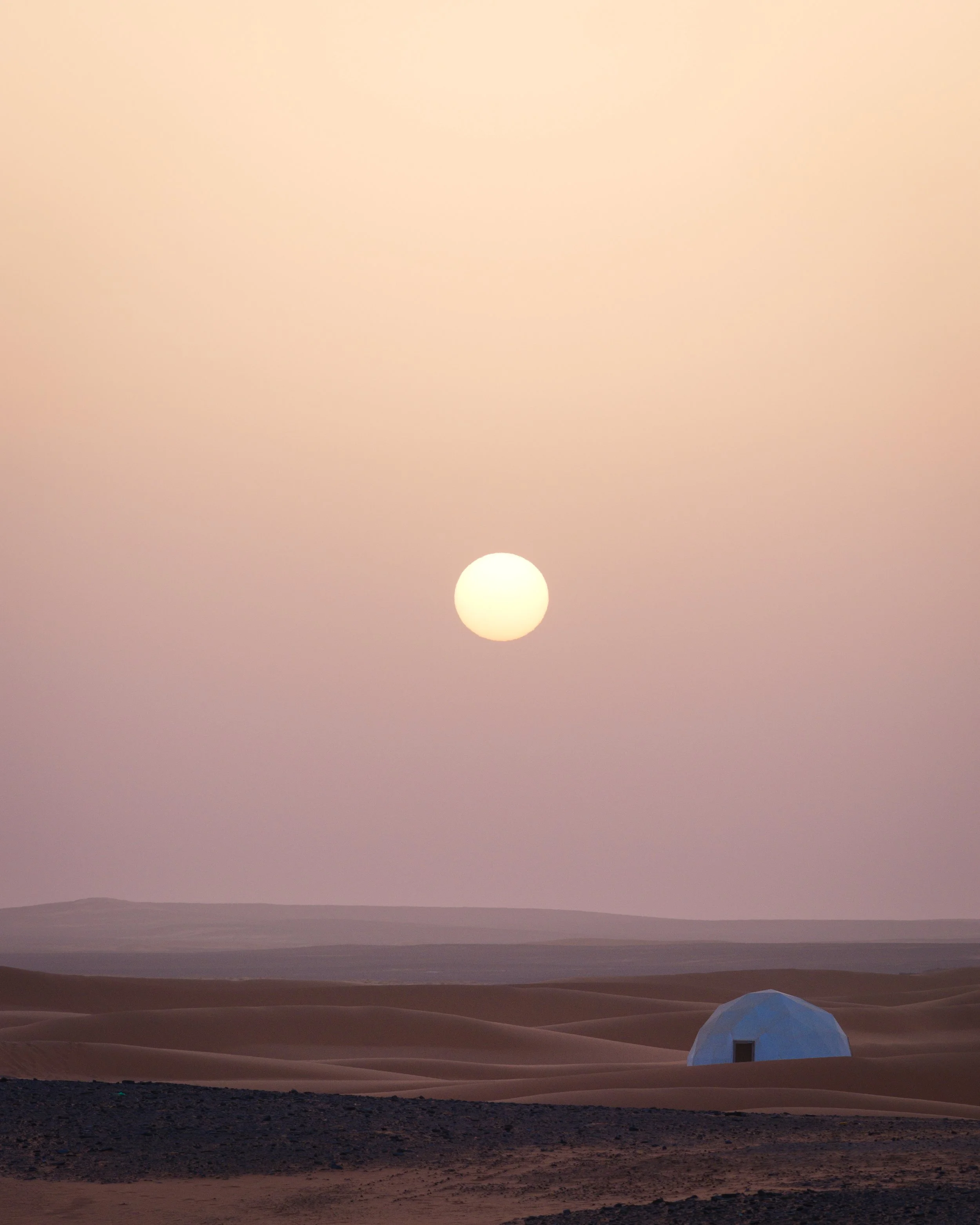 A desert landscape during sunset or sunrise with sand dunes, a small blue-domed structure, and the sun low in the sky.