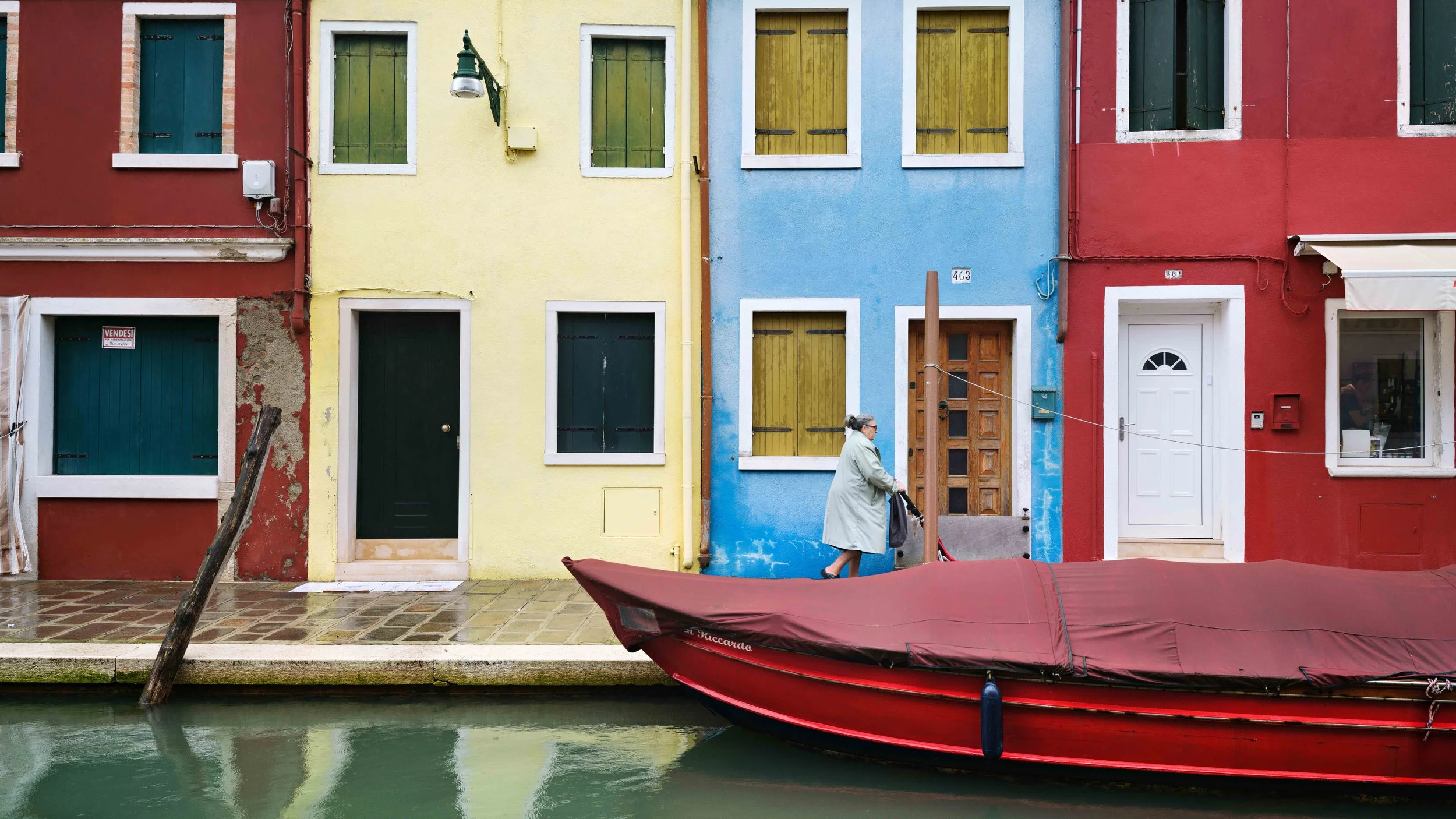 Colorful buildings lining a canal with a red boat docked in front and a woman walking on the sidewalk.