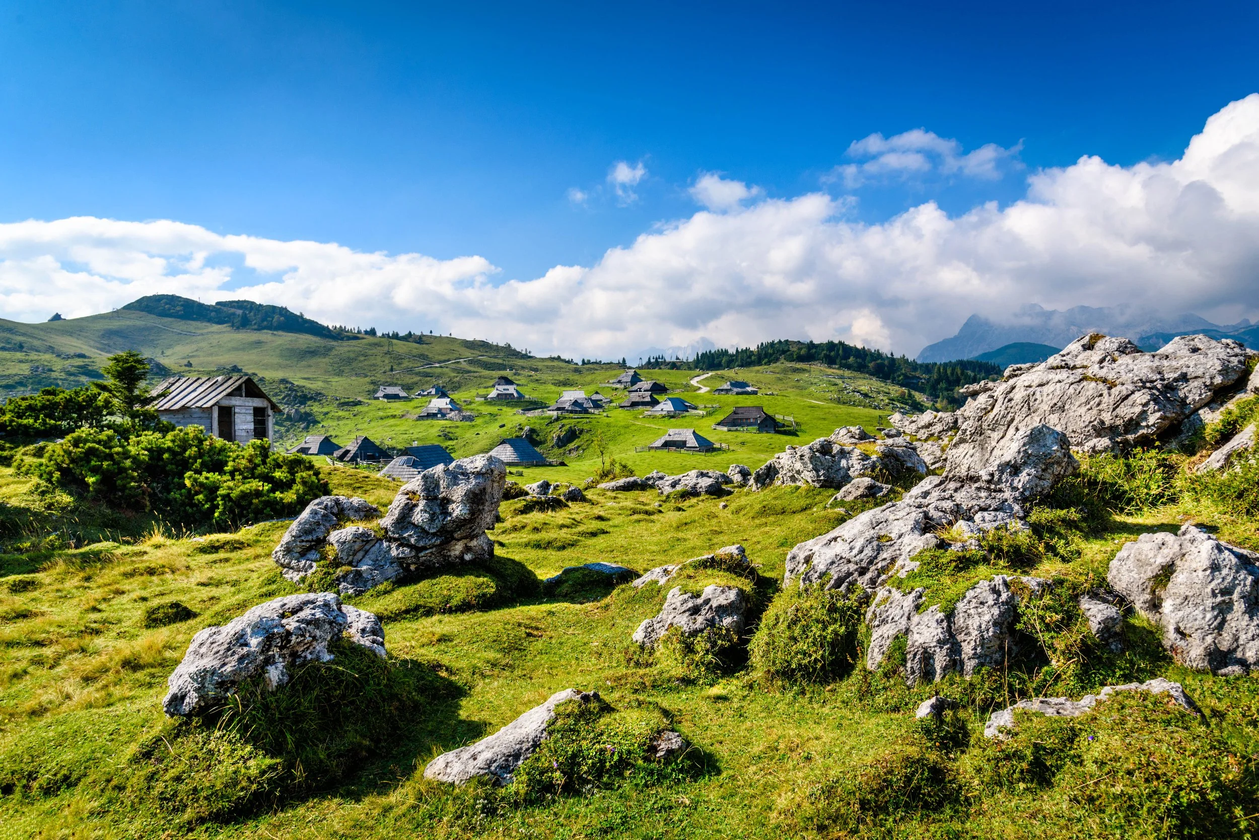 Rural landscape with green hills, scattered rocks, and small houses with blue roofs on a sunny day with a partly cloudy sky.