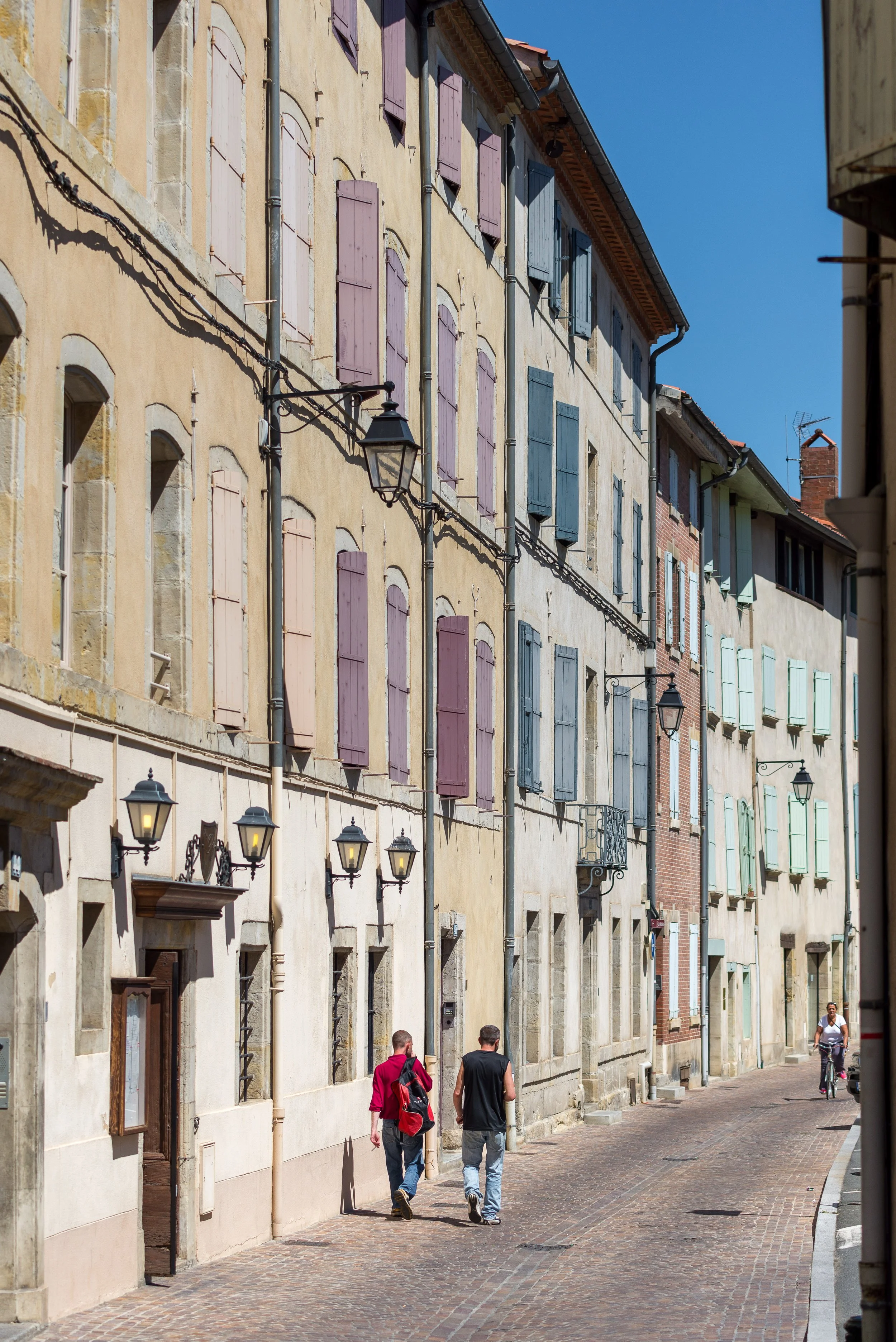 Colorful old European buildings with pastel shutters along a narrow cobblestone street, with two men walking and a cyclist in the background under a clear blue sky.