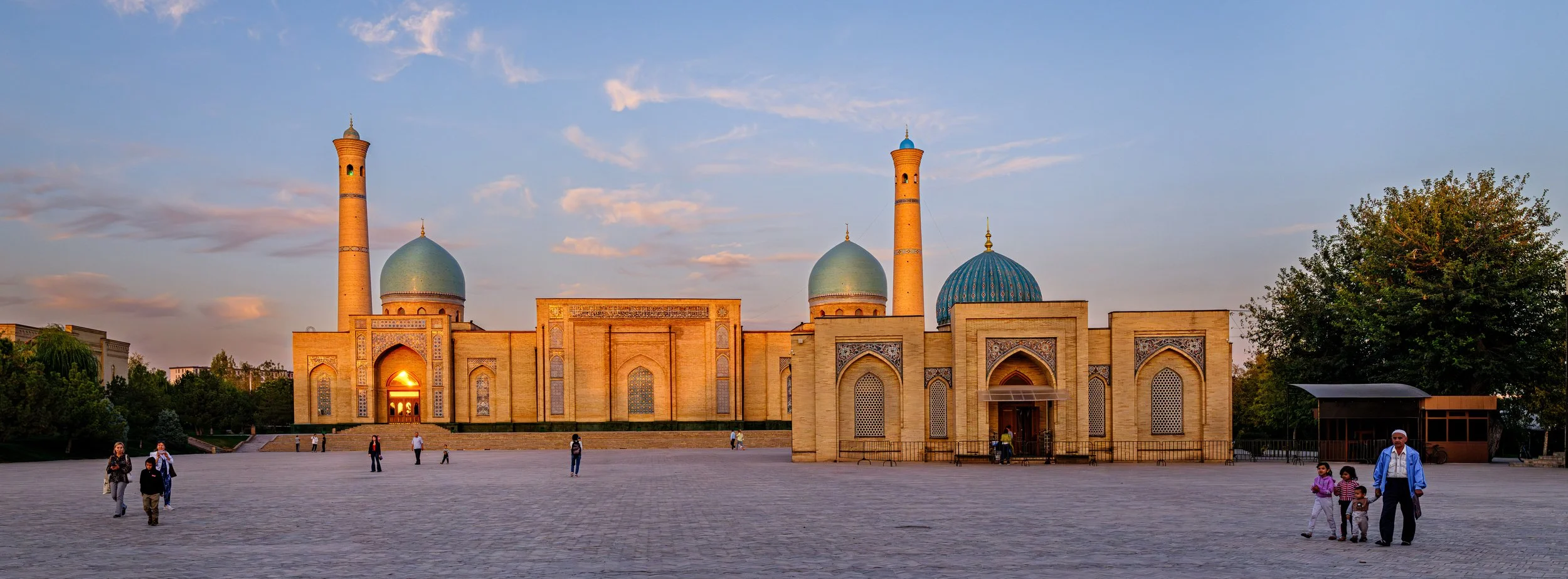 A mosque with blue domes and minarets, sunset sky, people walking in front.