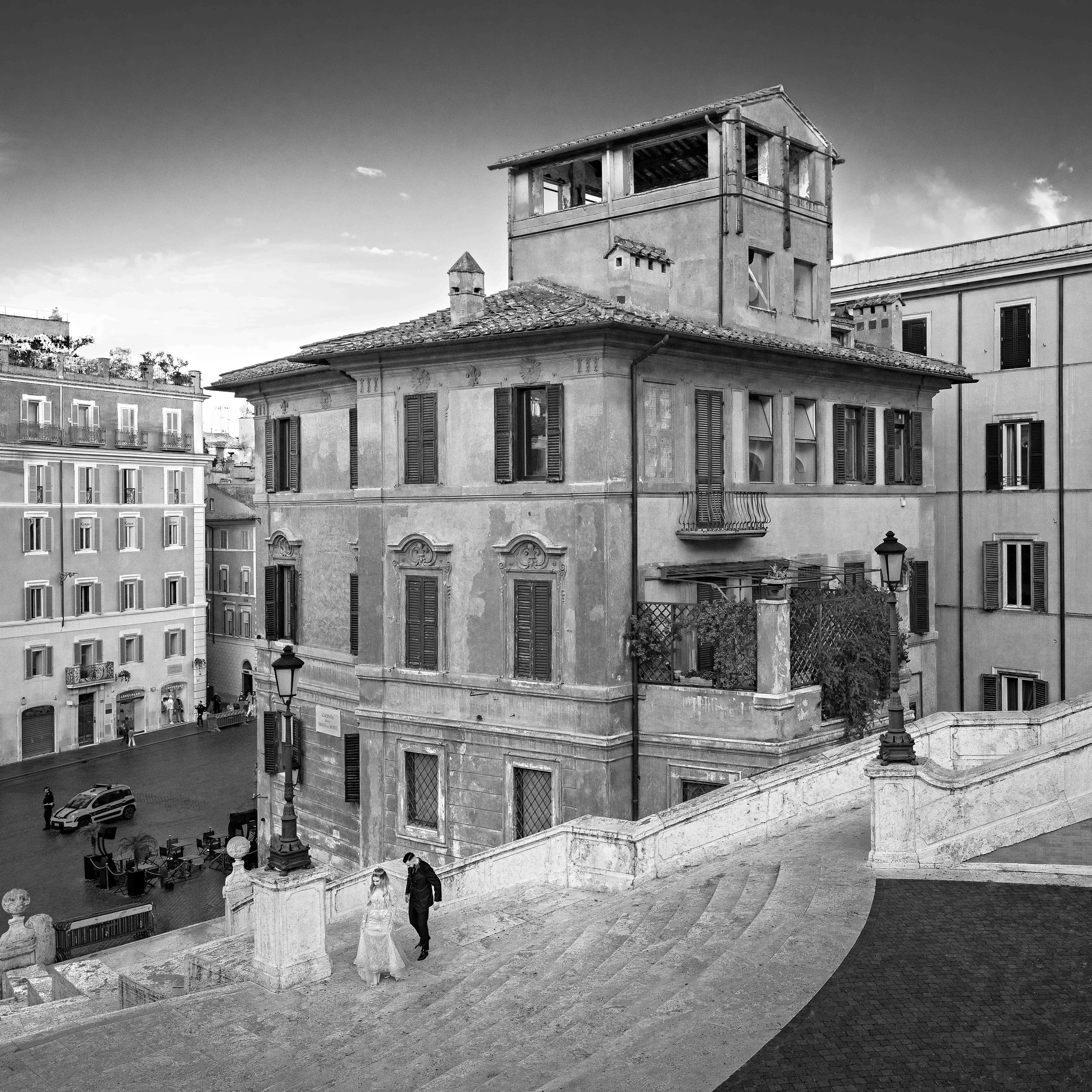 Black and white photo of an old building with shutters, a terrace, and a tower, alongside other buildings, with a street below and a couple walking up stairs.
