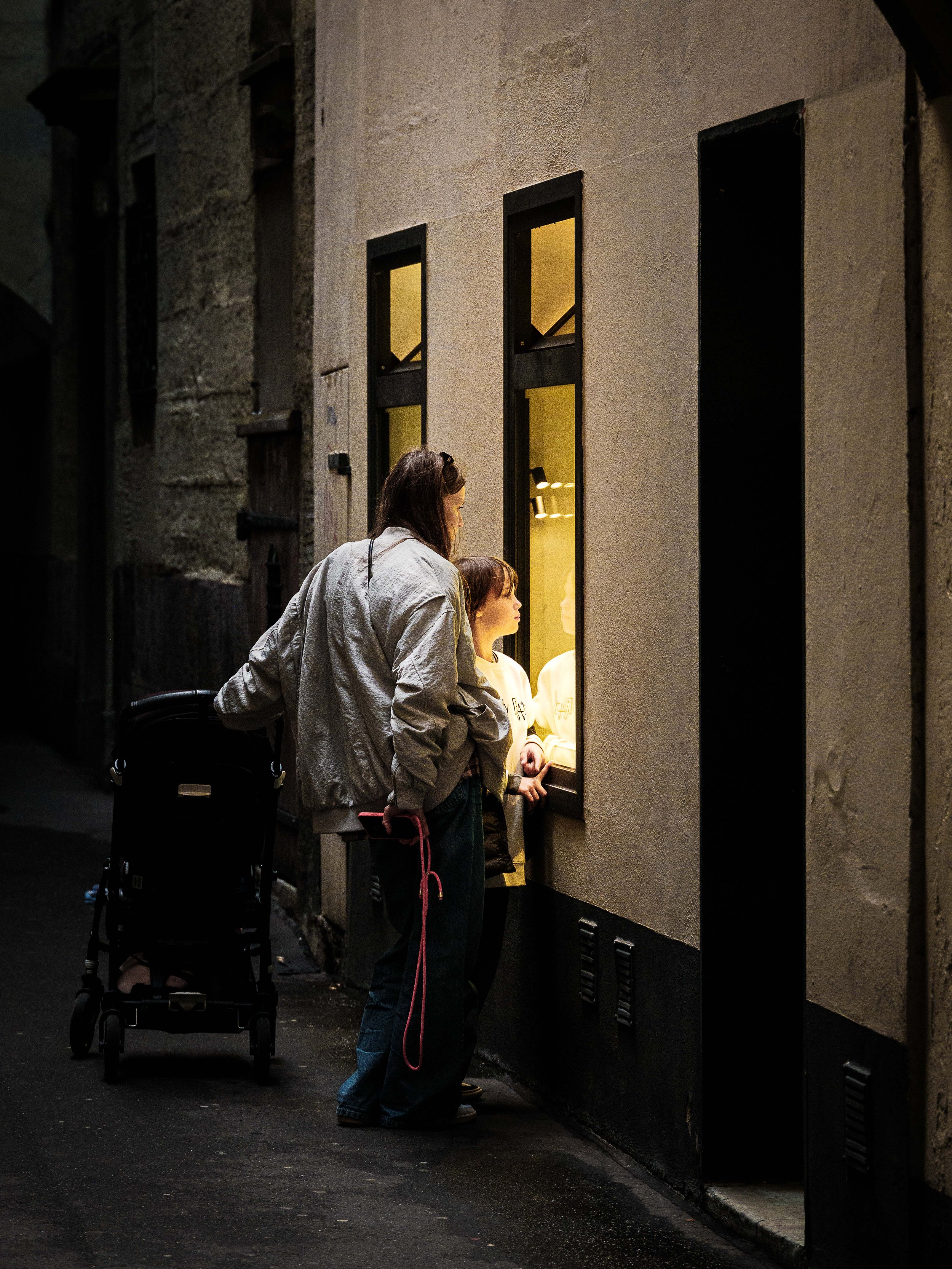 A woman and a girl looking through a window at night, with a stroller nearby.