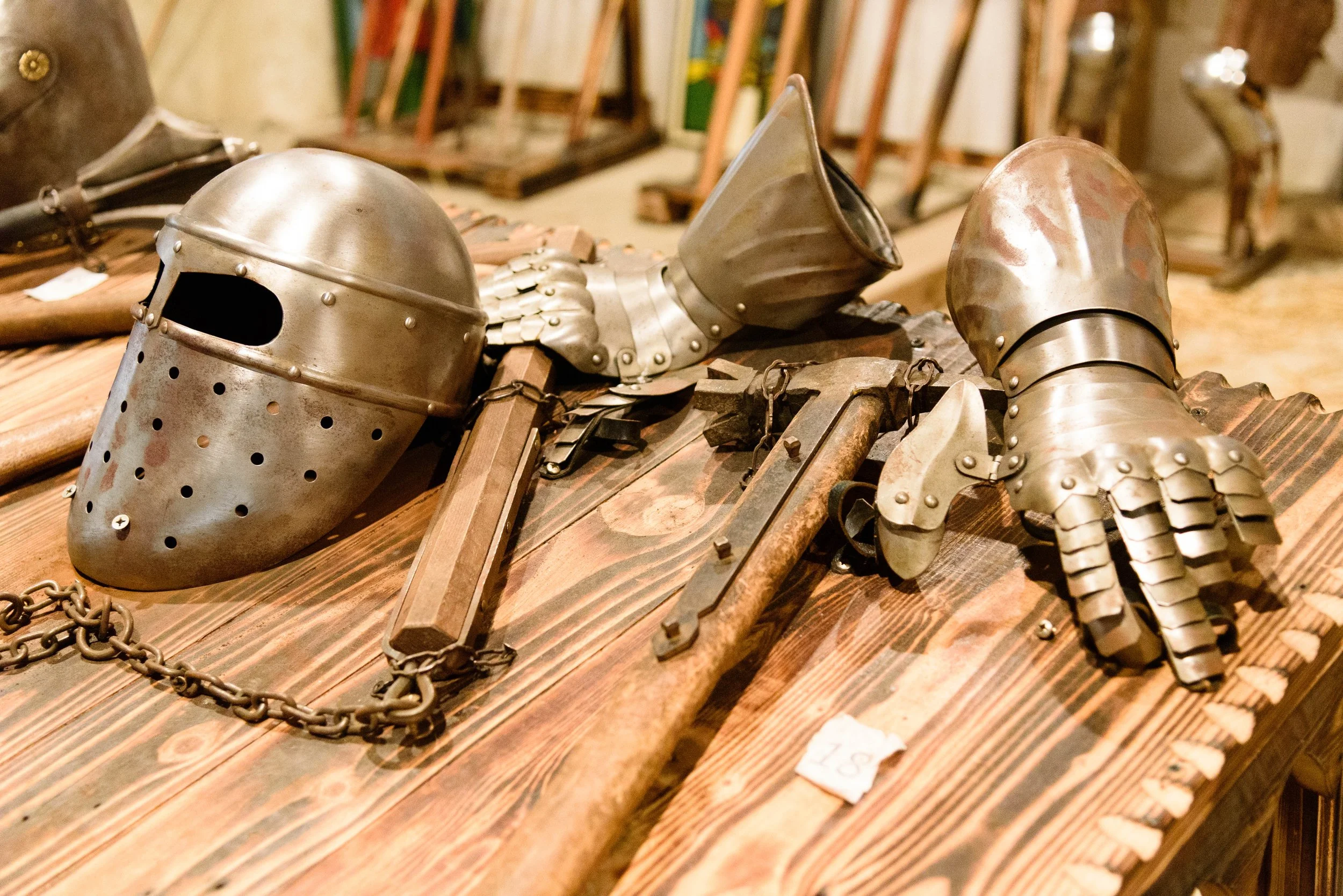 A collection of medieval armor and weapons displayed on a wooden table. Items include a metal helmet, a gauntlet, an axe, and a chainmail glove.