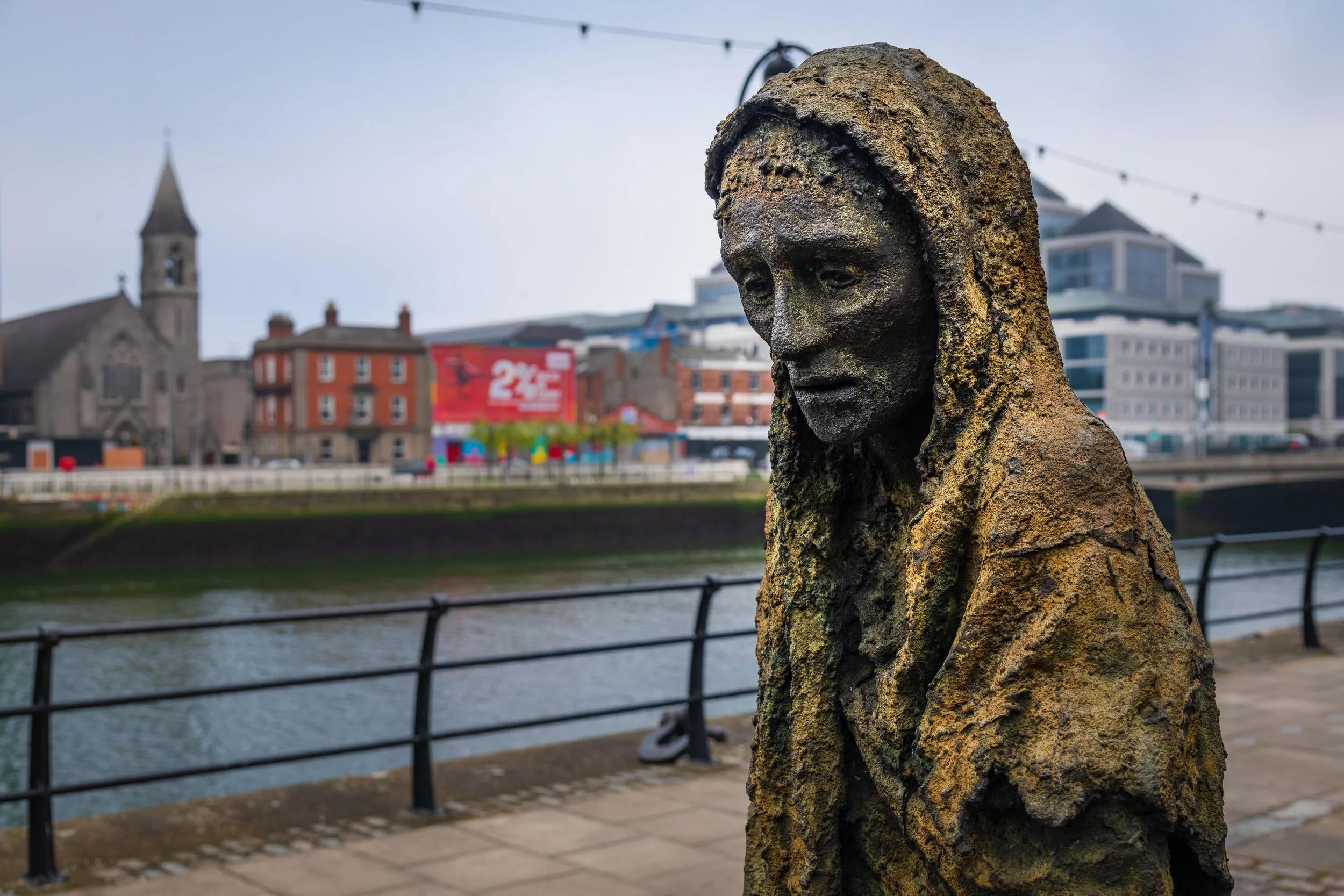 A weathered bronze statue of a person with a contemplative expression, by a river with city buildings and a church tower in the background.