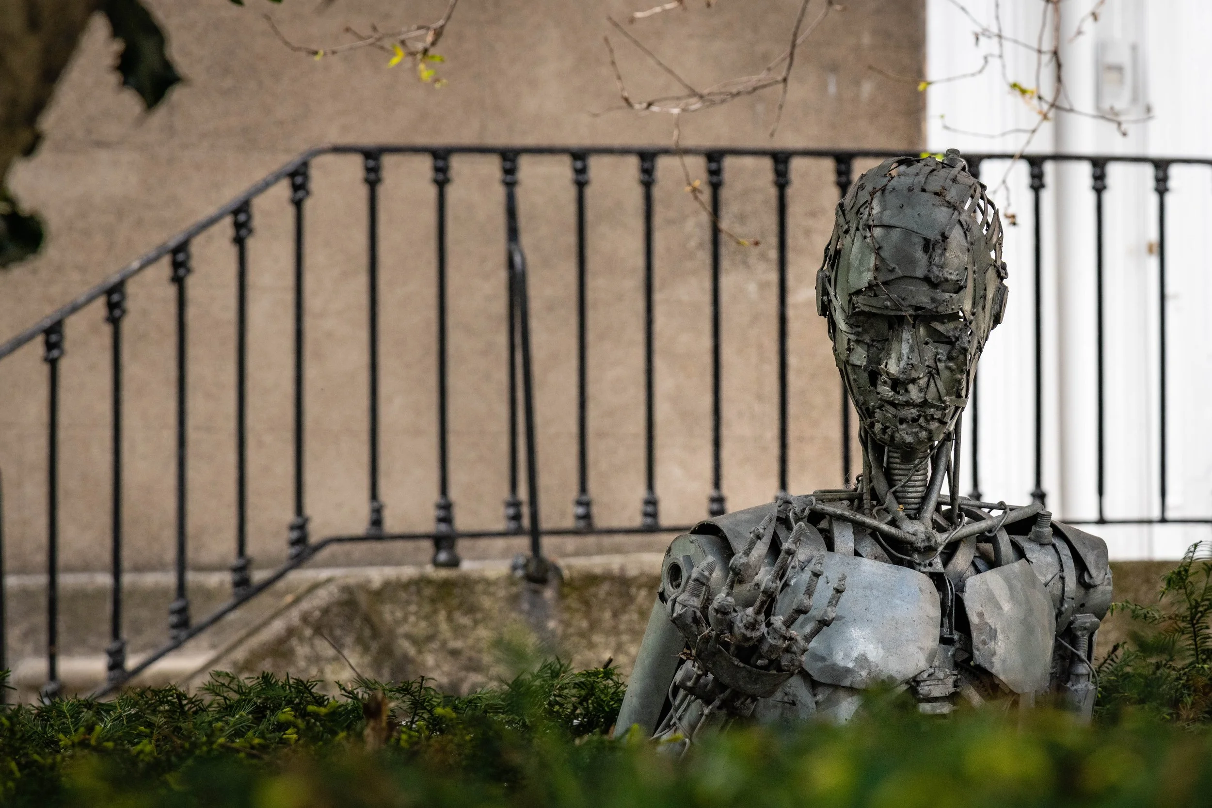 Sculpture of a human figure made from metal parts, sitting outdoors with a fence and stairs in the background.
