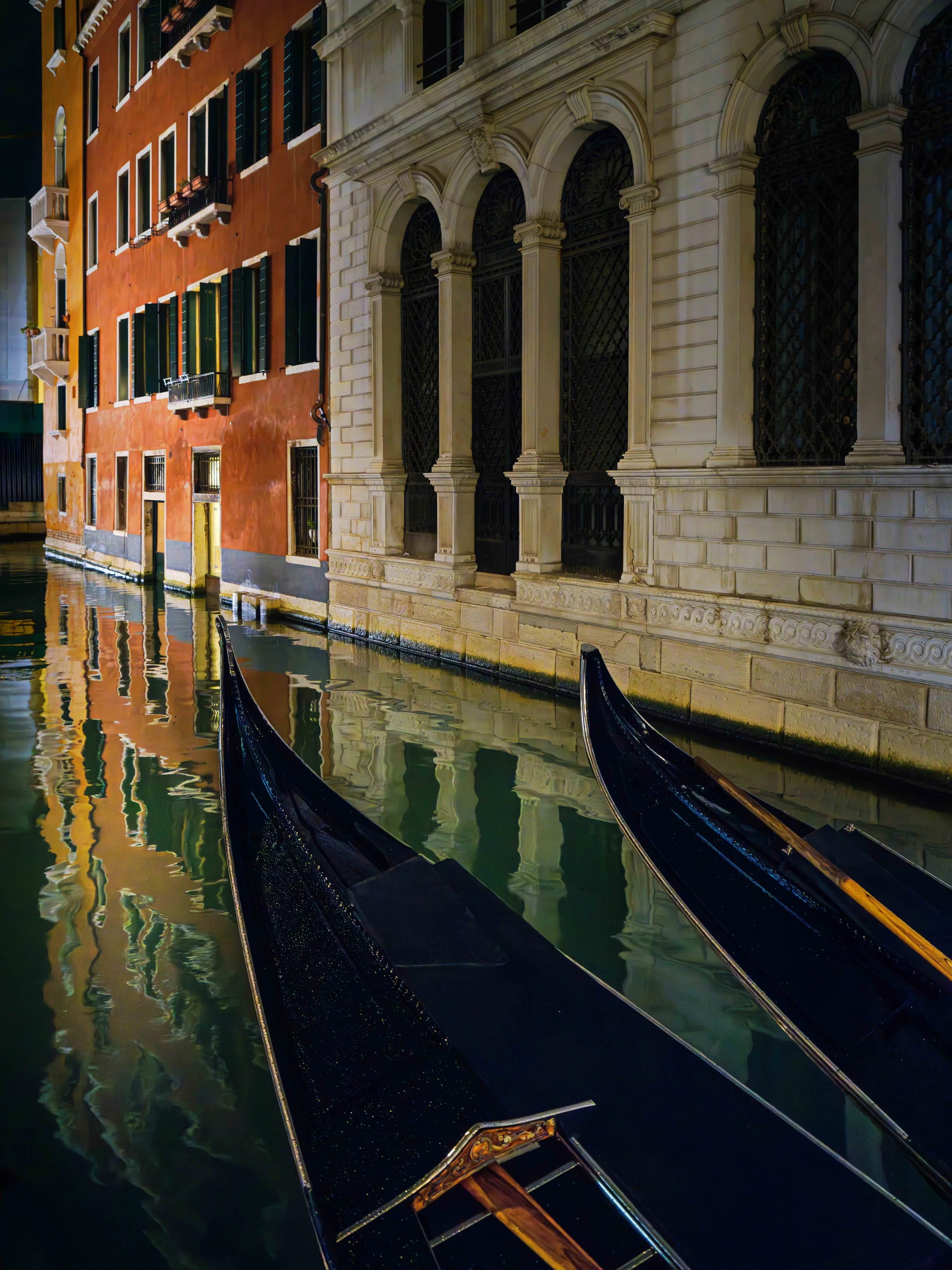 Nighttime scene of a narrow canal with two black gondolas and historic European-style buildings reflecting in the water.